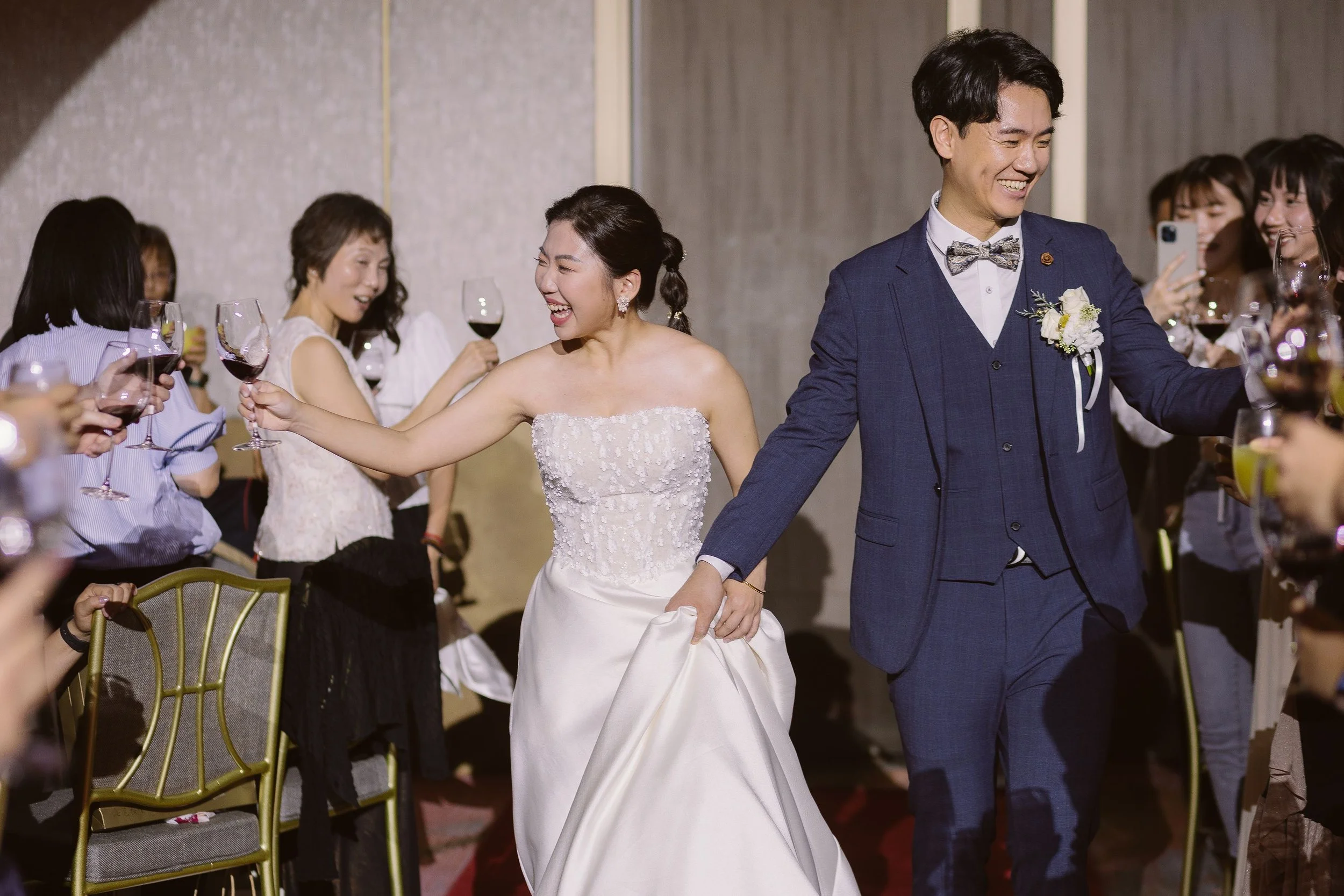 A bride and groom, both smiling, holding hands and toasting with wine glasses at their wedding reception surrounded by friends.