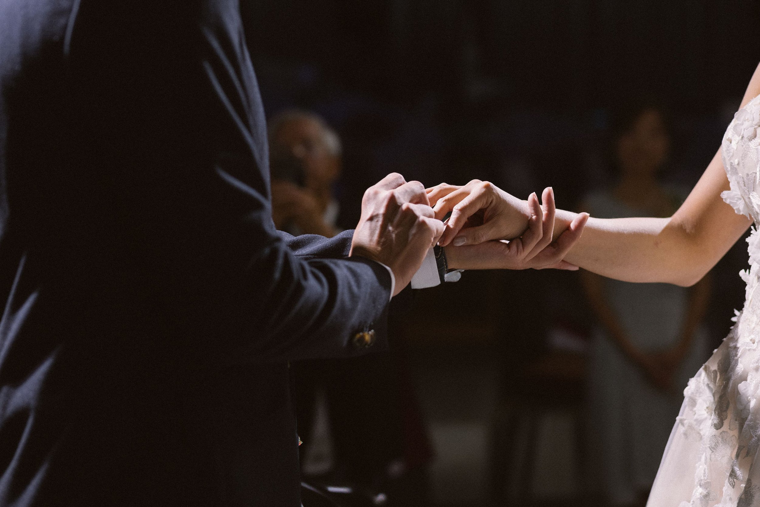 A couple holding hands during a wedding ceremony, with one person wearing a tuxedo and the other wearing a white lace dress.
