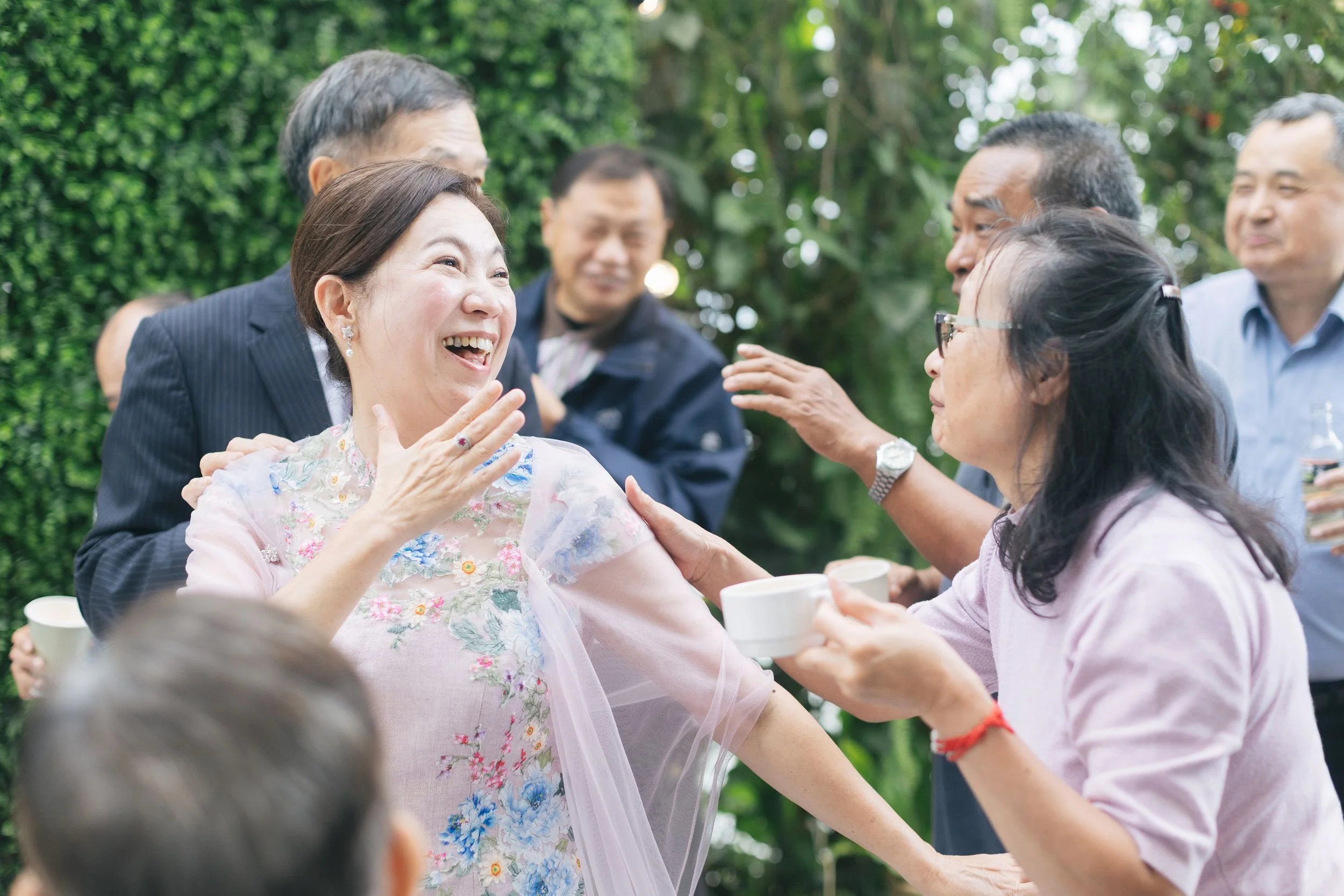 A group of people outdoors, with a woman in floral clothing smiling and engaging with others, some holding cups, surrounded by greenery.