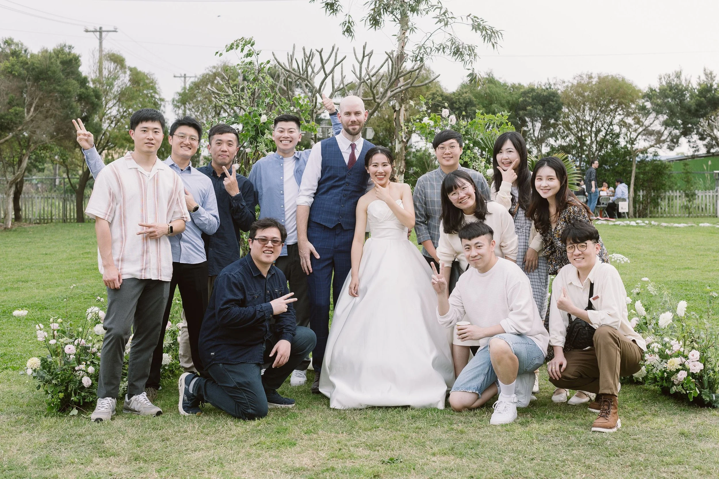 A group of people dressed in formal and casual attire gathering outdoors for a celebration, with some making peace signs, in front of a wedding couple in a garden setting.