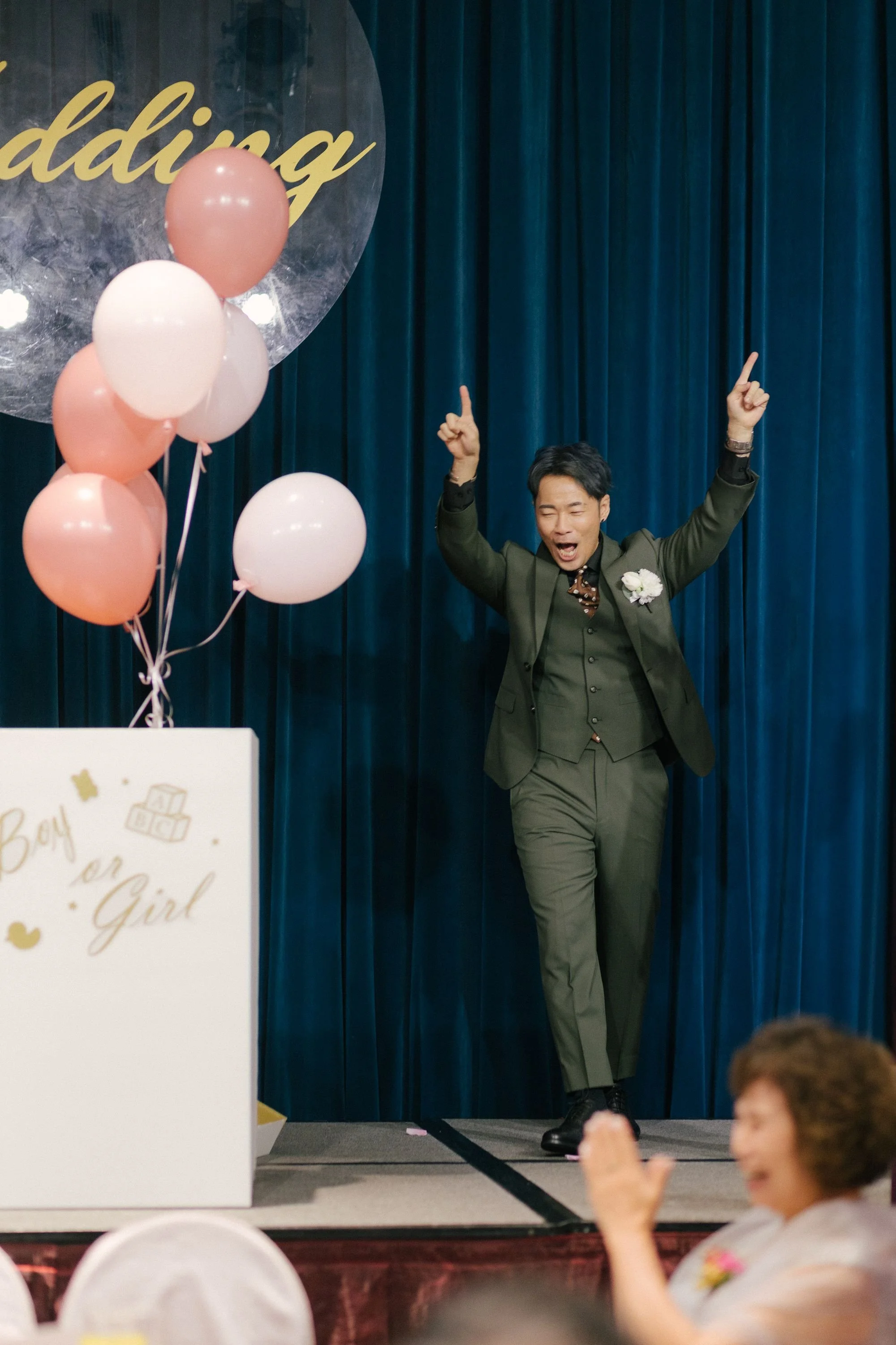 A man in a suit with a flower boutonniere is celebrating on stage with his arms raised. There are pink and white balloons nearby, and a decorated sign that says 'Baby on Girl' partially visible. The background features dark blue curtains, indicating 