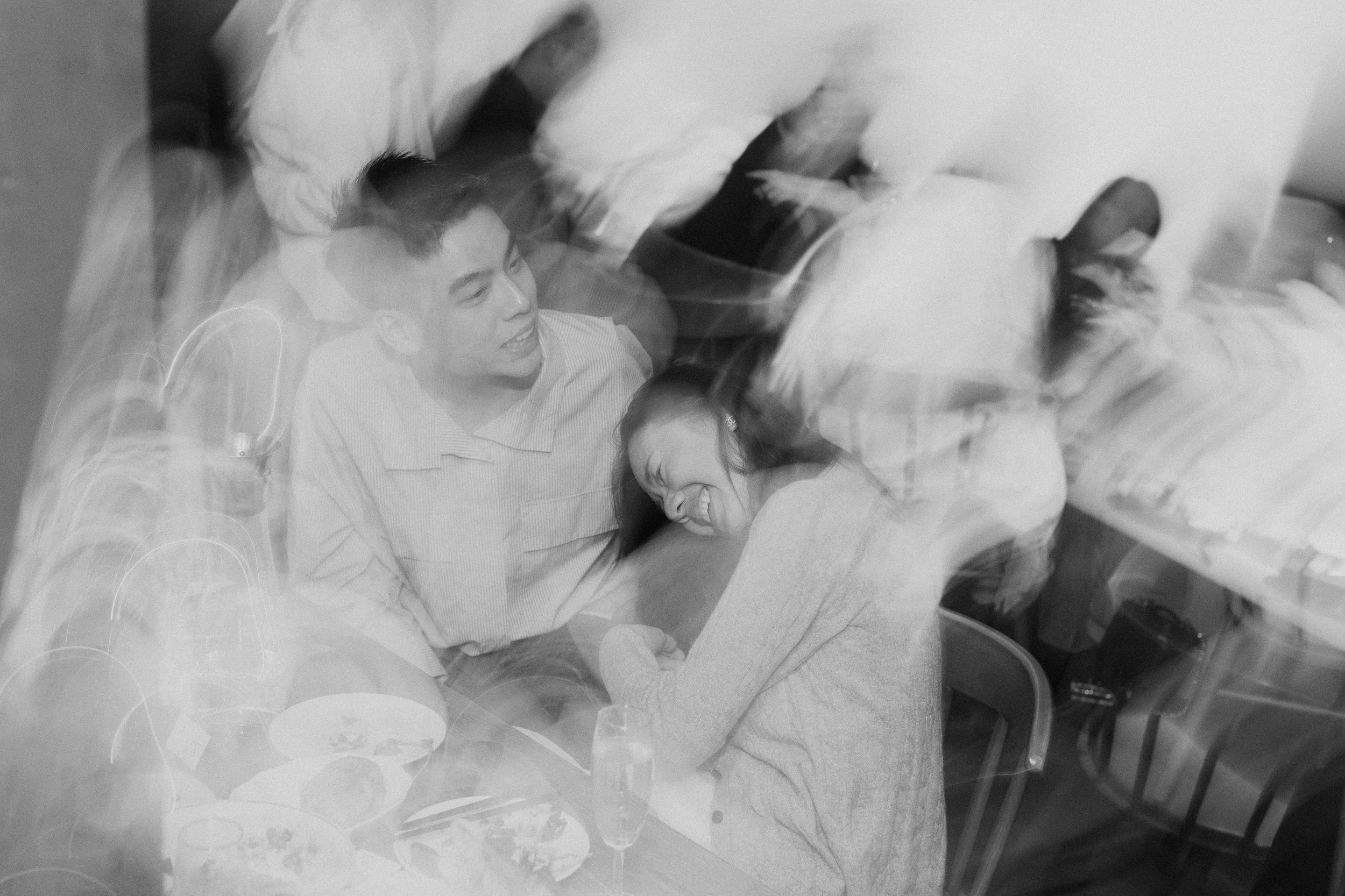 Black and white photo of two women smiling and lying on a table or bed, with one woman's head resting on the other's shoulder. The image has a dreamy, double exposure effect.