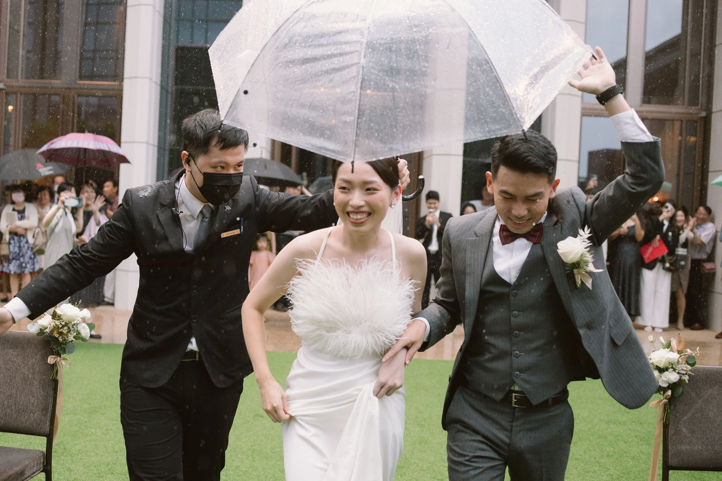 A bride in a white wedding dress smiling, holding her dress, walking with two men in suits, one pointing out and the other holding an umbrella, on a rainy day at a wedding reception with guests in the background.