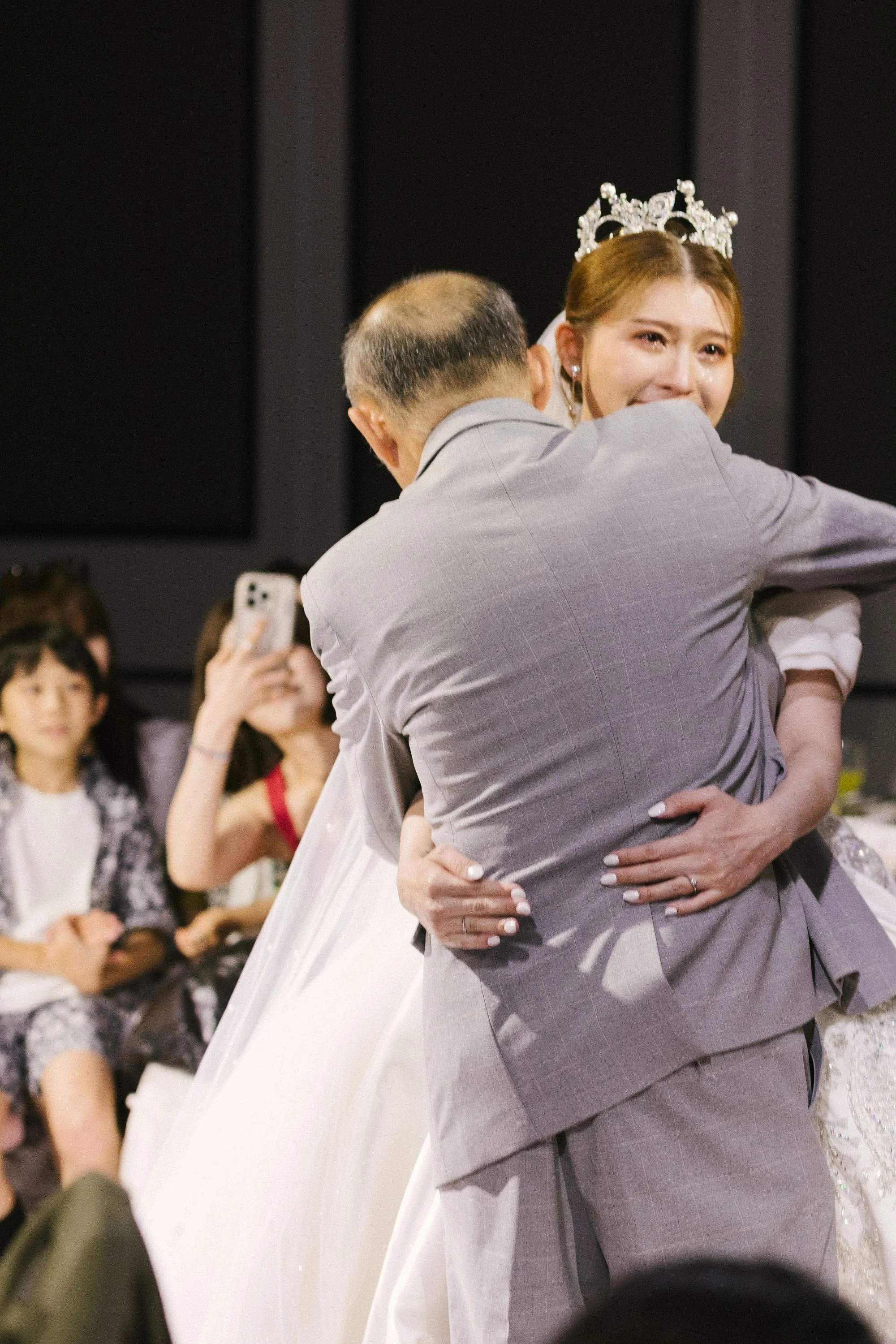 A woman with a tiara and makeup, crying tears of joy, being embraced by an older man at a celebration or event, surrounded by onlookers taking photos.
