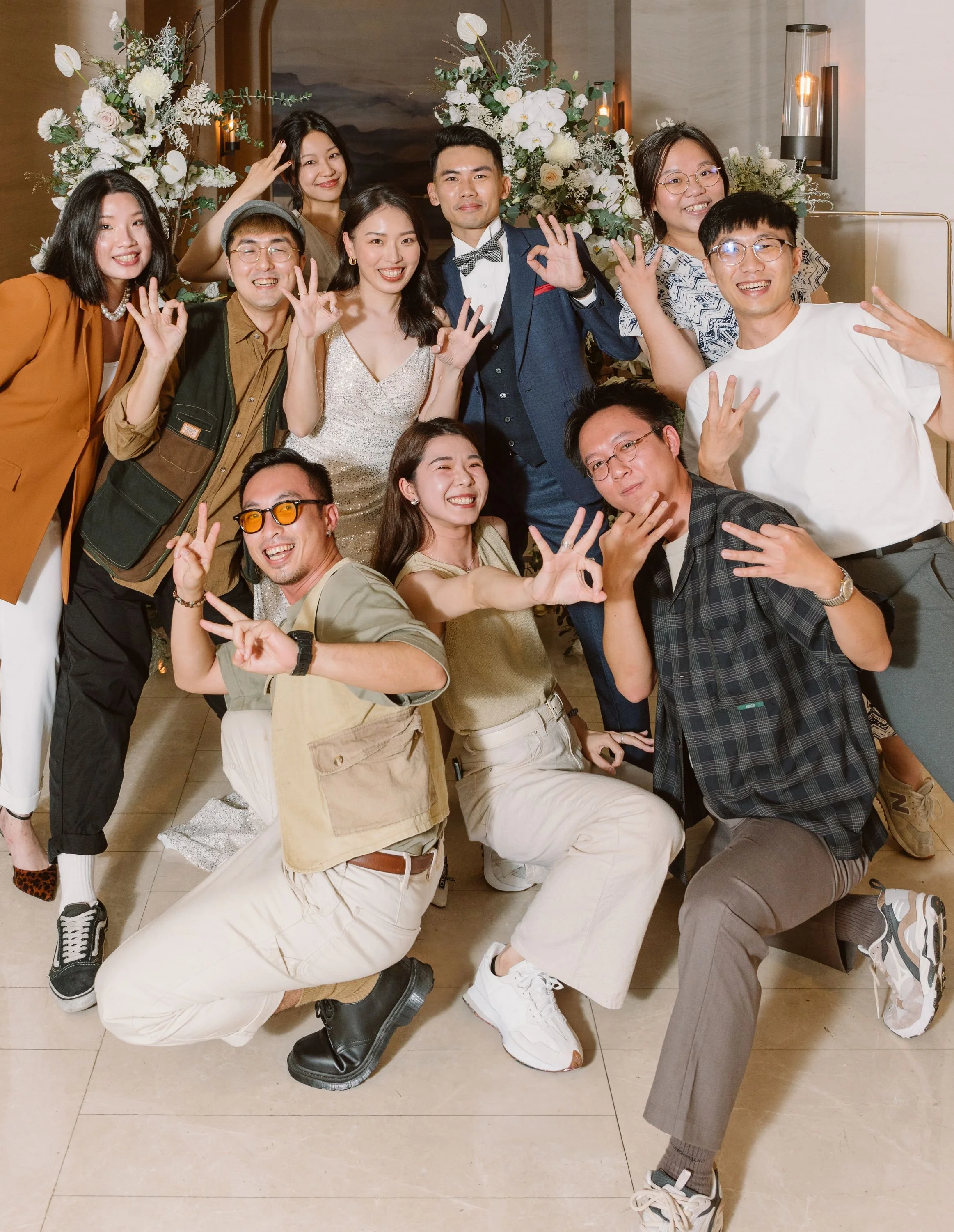 A group of people in formal attire celebrating at a party with floral decorations in the background, smiling and making peace signs.