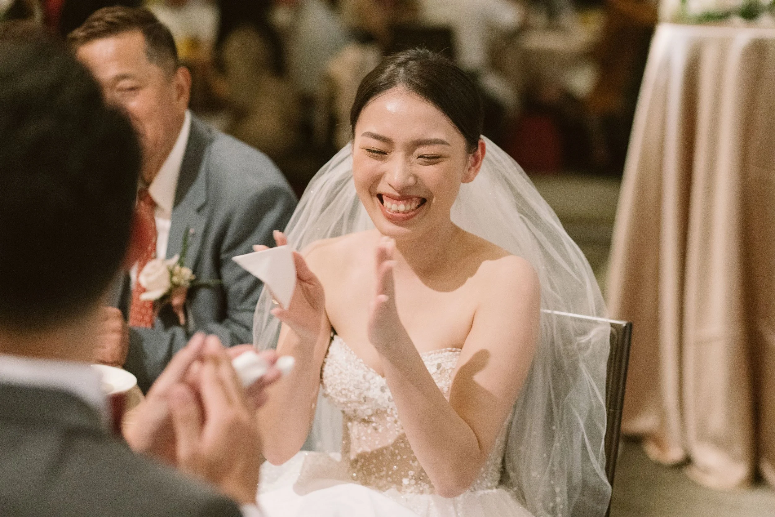 A bride with dark hair styled back, smiling and looking at a guest, during a wedding reception. She is wearing a white wedding dress and veil, and is seated at a table with guests.