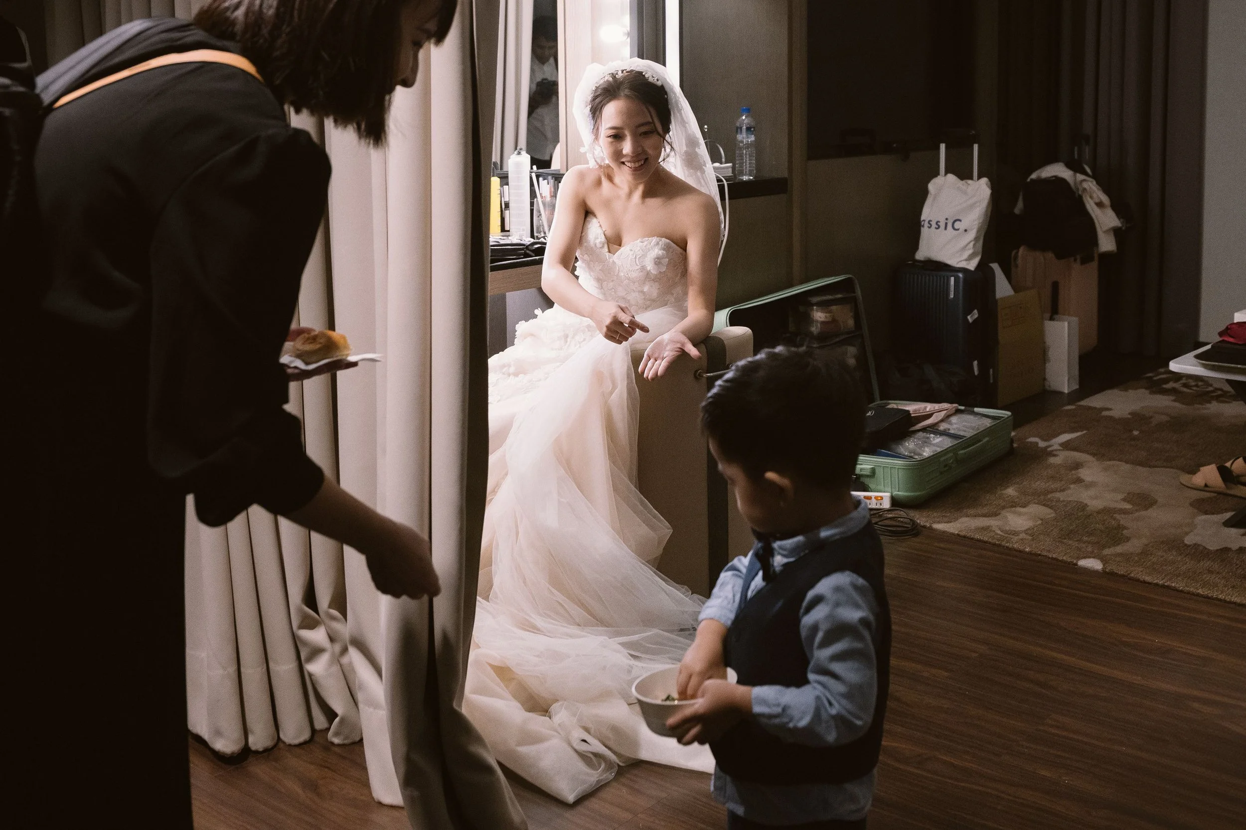 A bride in a white wedding dress smiling and sitting on a chair, surrounded by children and an adult, in a room with a dresser, mirror, and luggage in the background.
