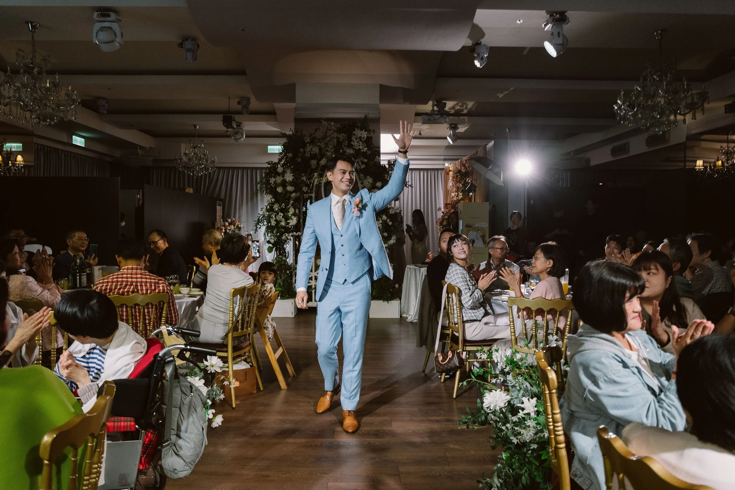 A man in a light blue suit walking and waving at a seated audience in a banquet hall decorated with flowers, with chandeliers on the ceiling and people applauding.