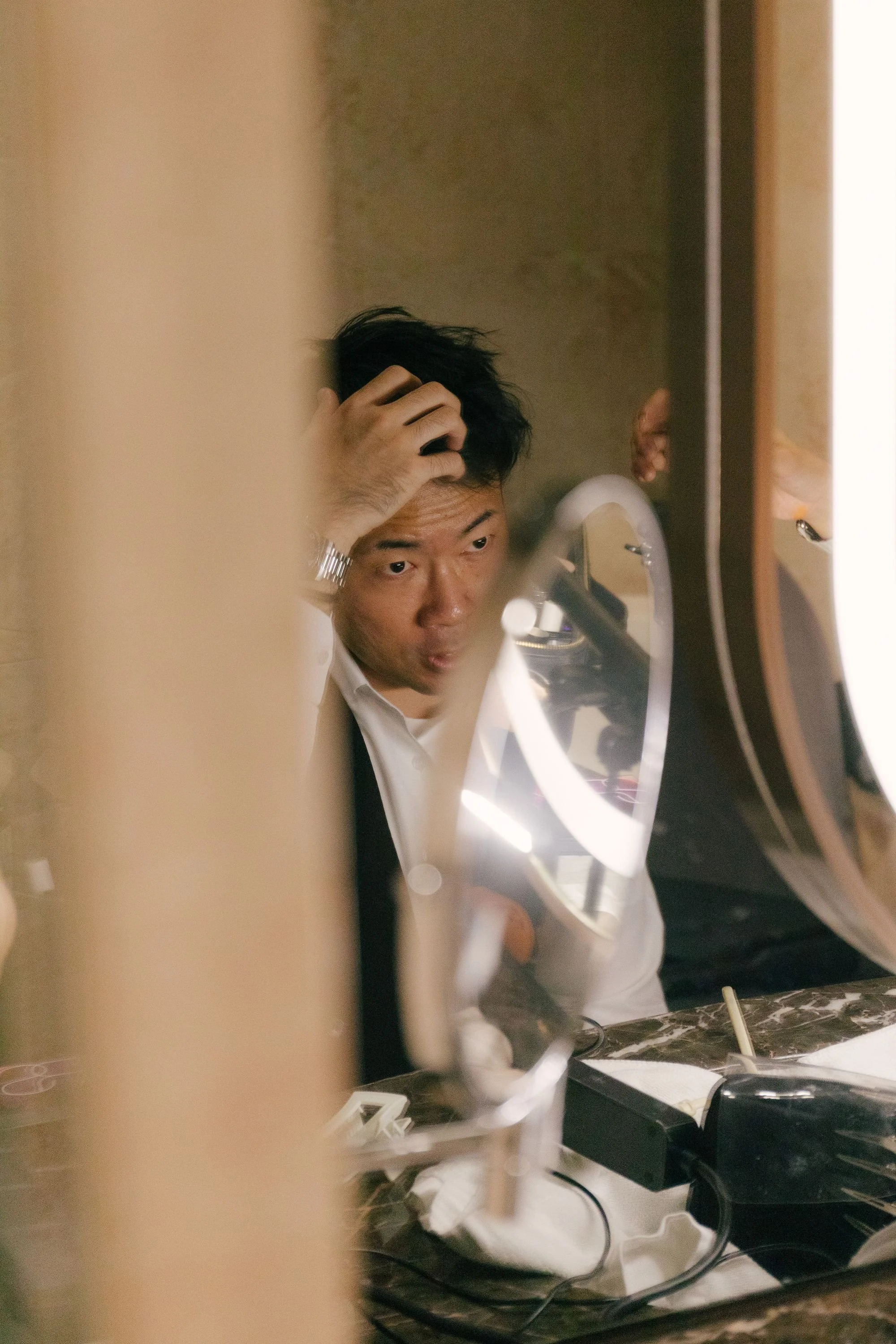 A man with dark hair, wearing a white shirt, is looking into a mirror while touching his head. The image is taken from behind a partial object, with a dark countertop and various cords and objects on it. The man appears to be in a bathroom or groomin