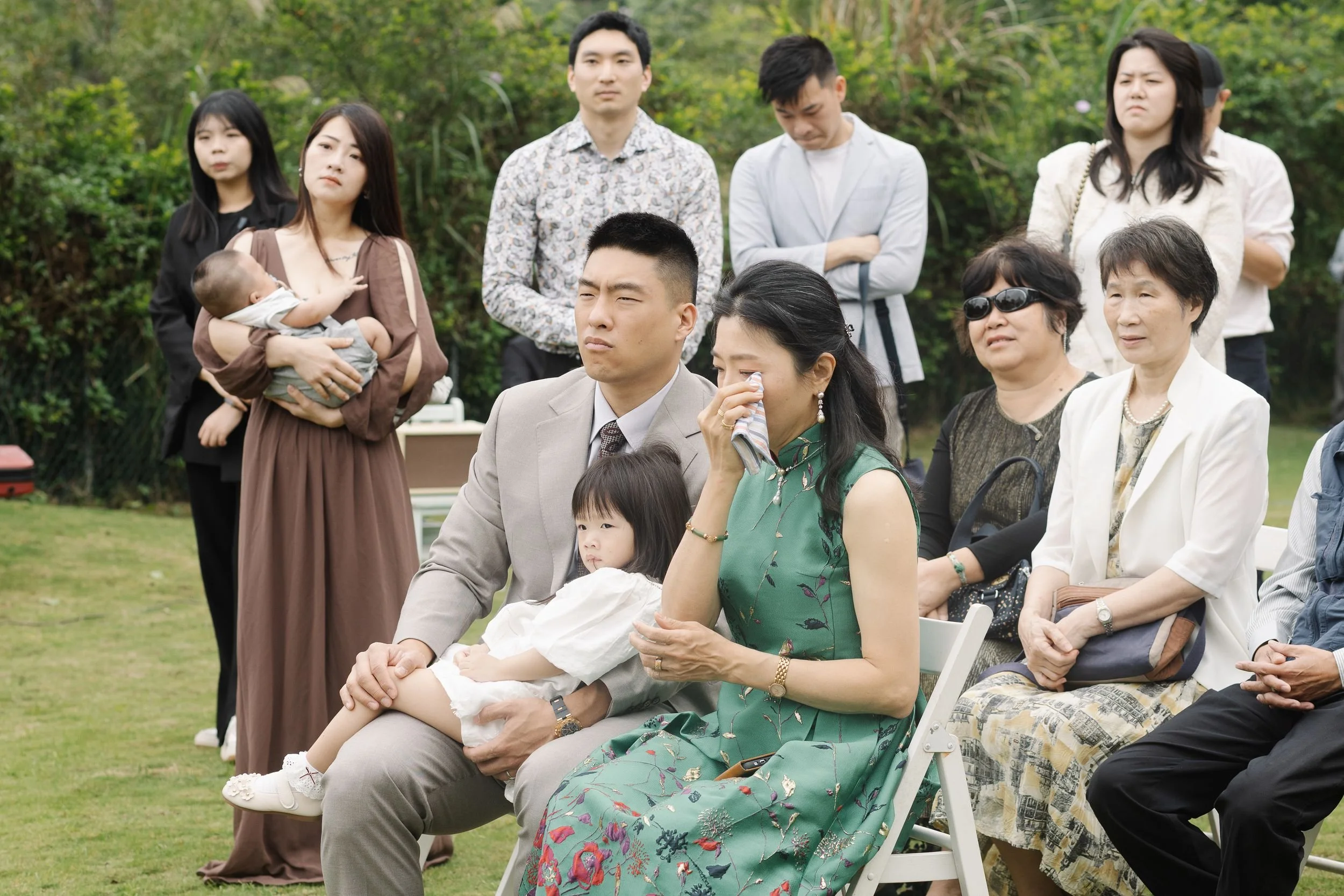 A group of people attending an outdoor event, with a woman in a green dress crying and a man holding a young girl on his lap. Others are standing or sitting around, some with serious expressions, amidst greenery.