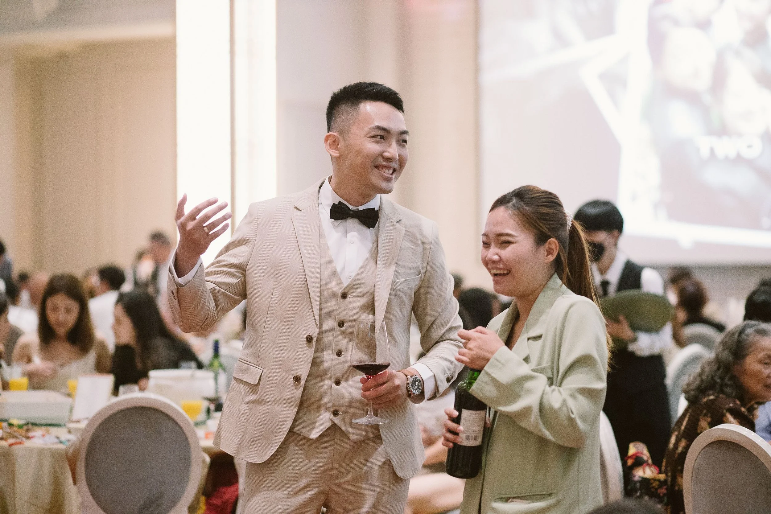A man in a beige suit and black bow tie talking to a woman in a light-colored blazer at a formal event, with other guests seated at tables in the background.