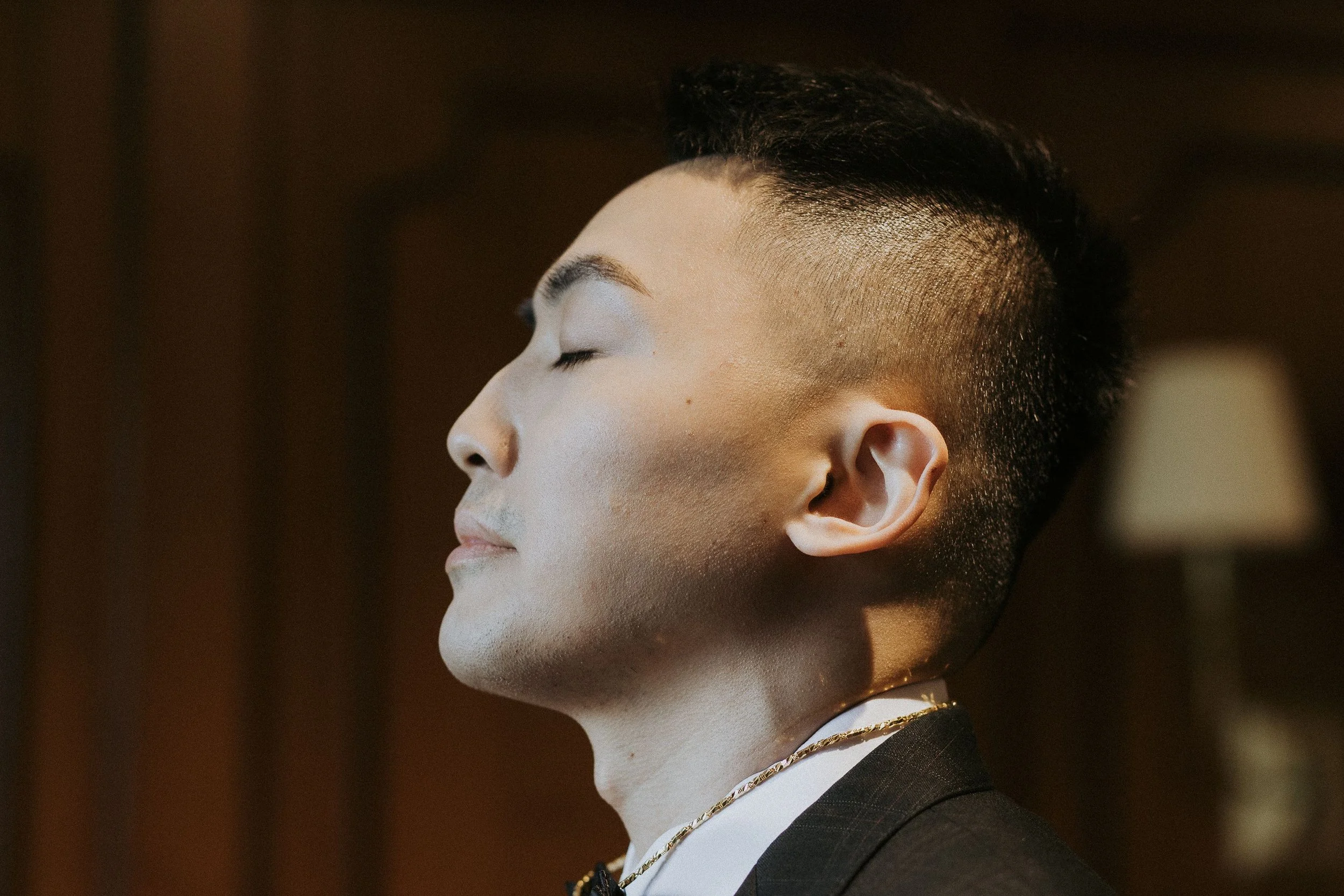 Side profile of a person with short, dark, textured hair, wearing a dark blazer, white shirt, and layered gold necklaces, with eyes closed and a neutral expression.