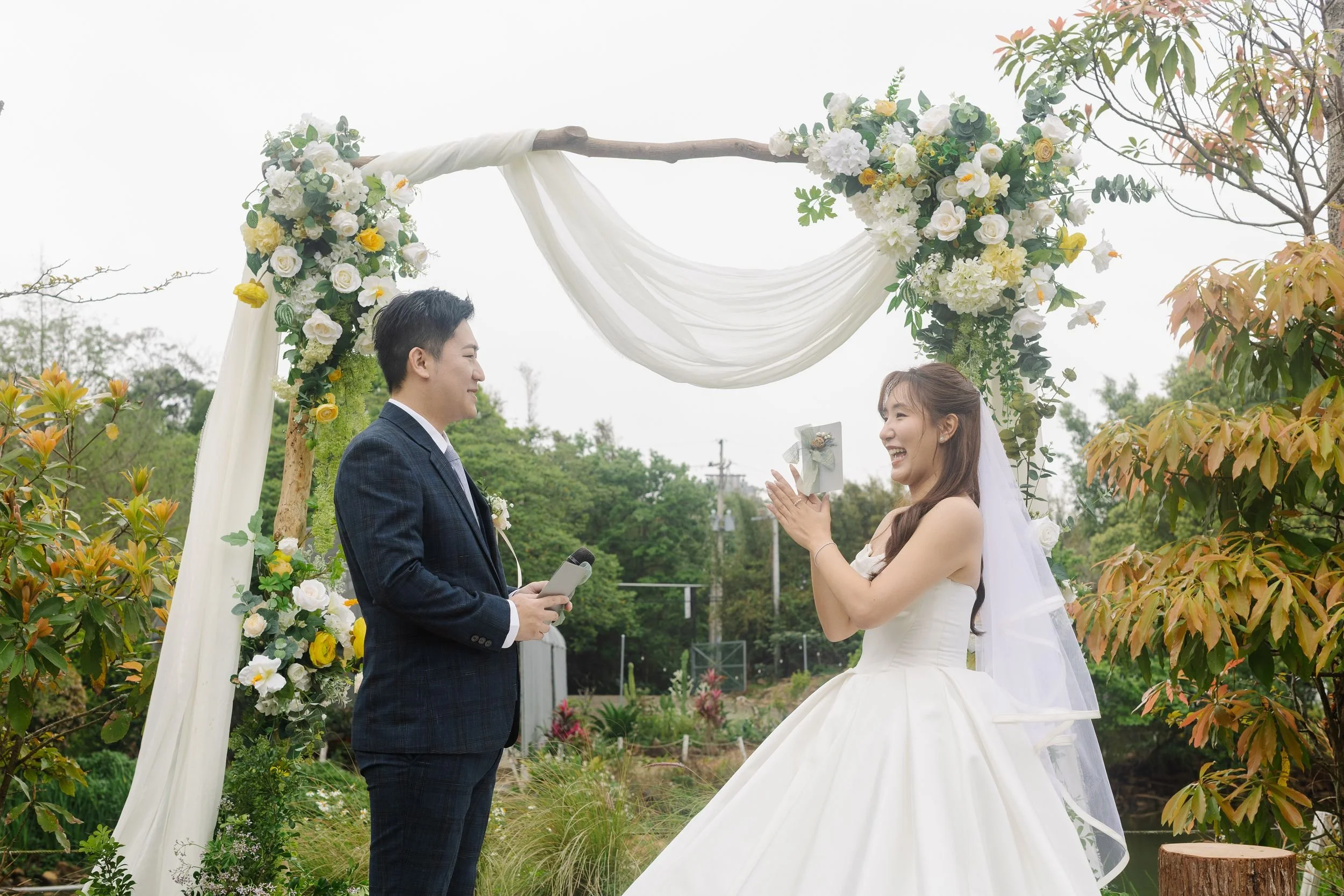 A bride and groom exchanging vows outdoors during a wedding ceremony under a floral arch, with trees and greenery in the background.