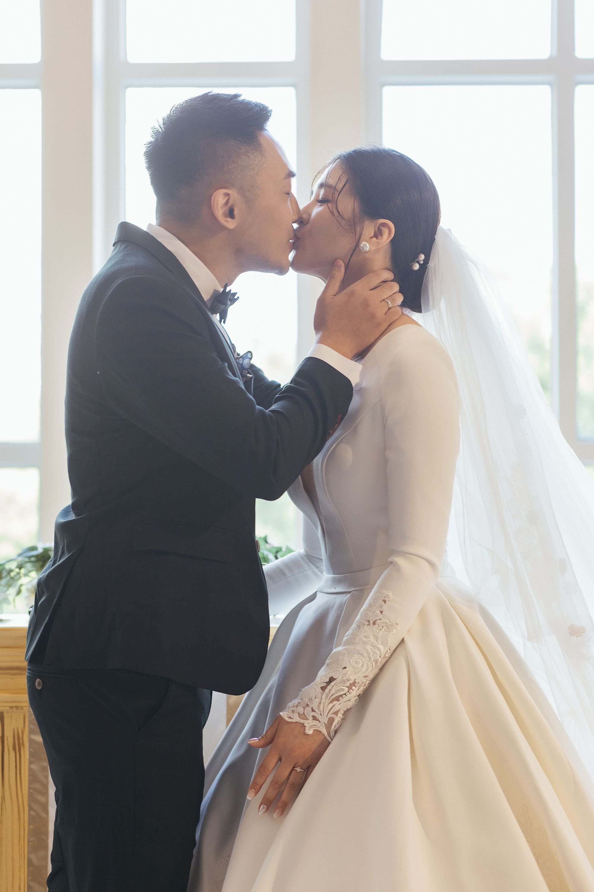 A newlywed couple sharing a kiss indoors with large windows in the background. The groom is wearing a black tuxedo, and the bride is in a white wedding dress with lace details at the sleeve. The bride has dark hair with pearl accessories and a veil.