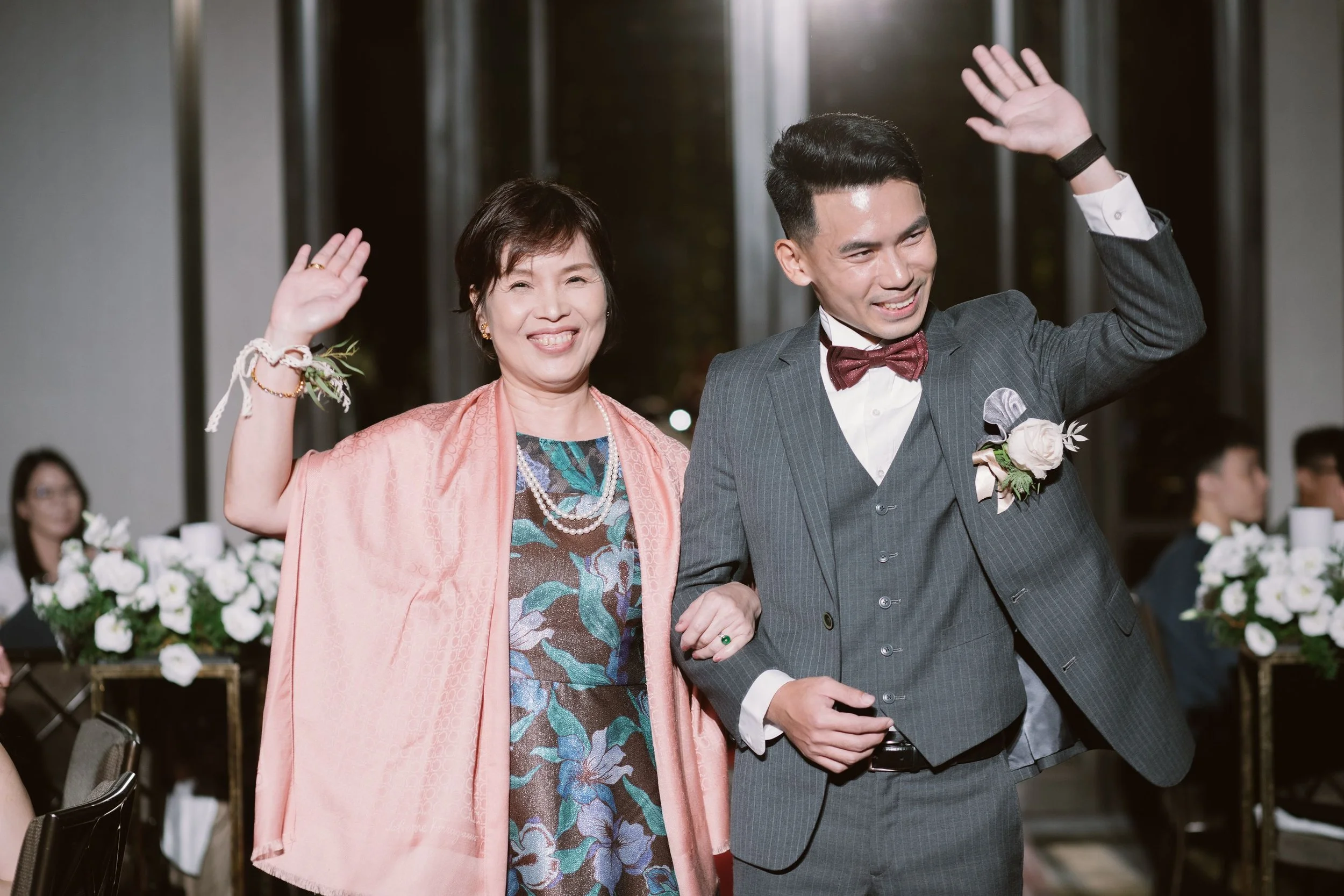 A joyful woman and a man dressed in a suit and bow tie are waving at a celebration or wedding, holding hands and smiling.