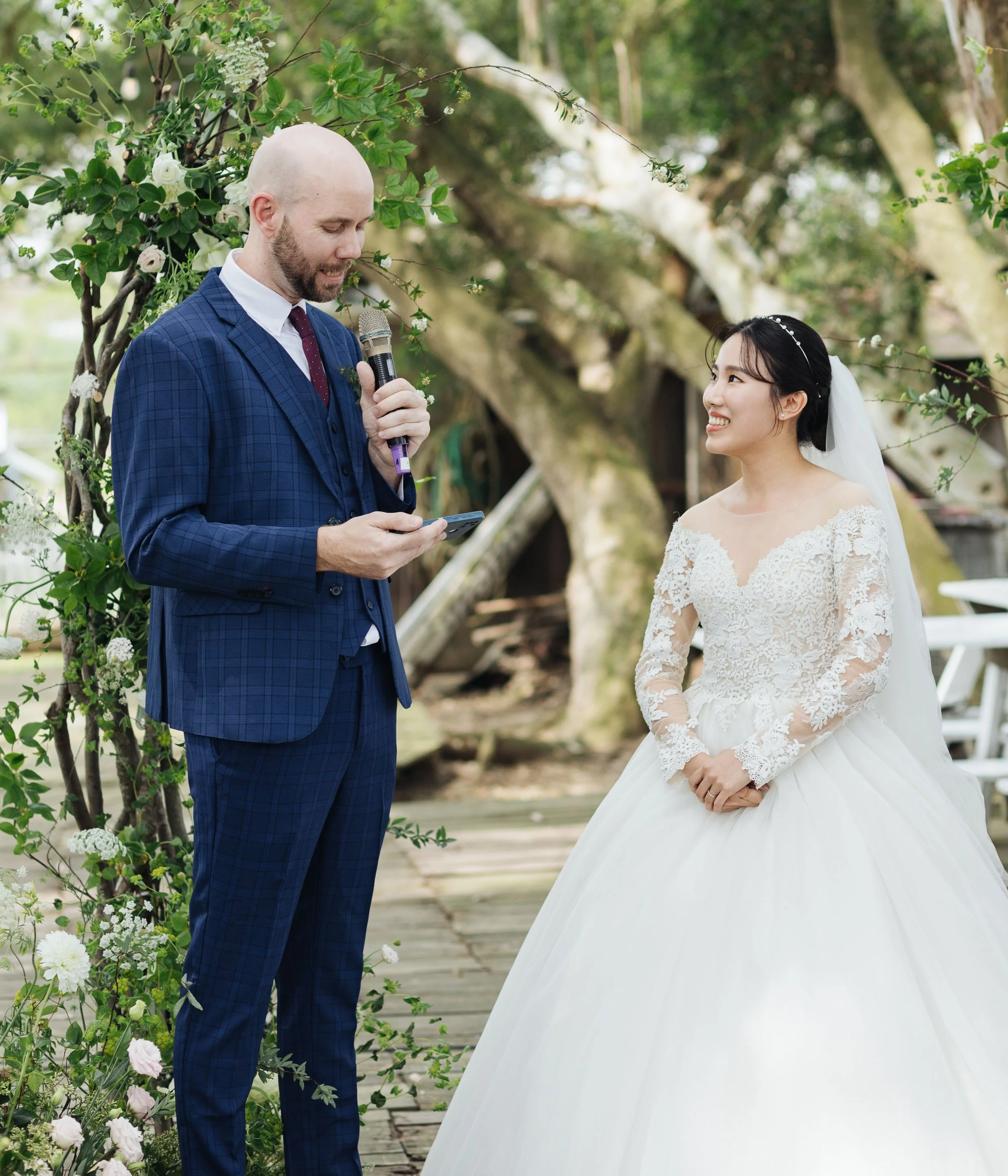 A groom with a bald head and beard wearing a blue checkered suit and a bride with black hair in an updo and wearing a white lace wedding dress, standing outdoors under trees, with the groom speaking into a microphone and the bride smiling at him.