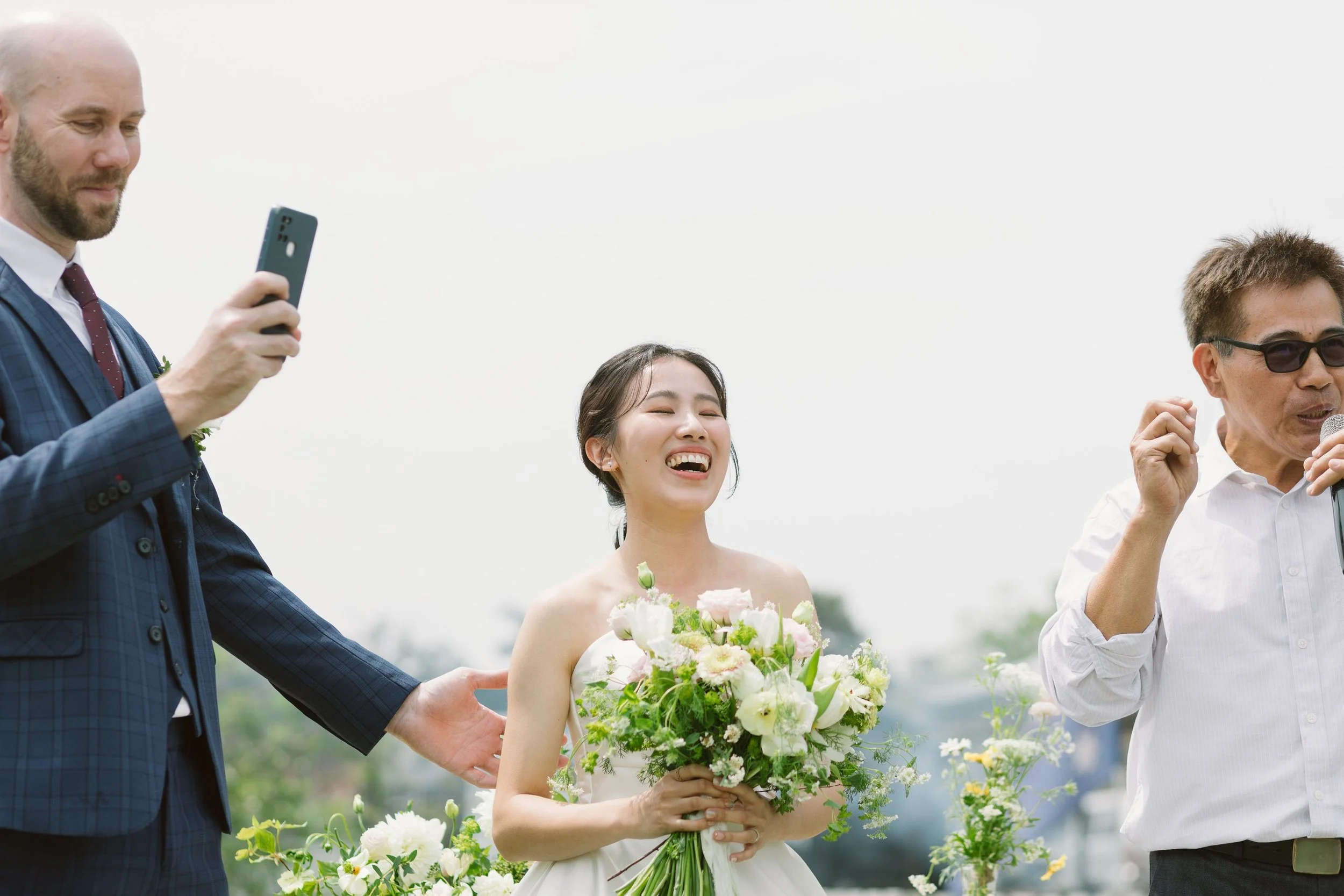 A woman in a wedding dress holding a bouquet of flowers, smiling, standing outdoors with two men, one taking a photo and the other speaking into a microphone.
