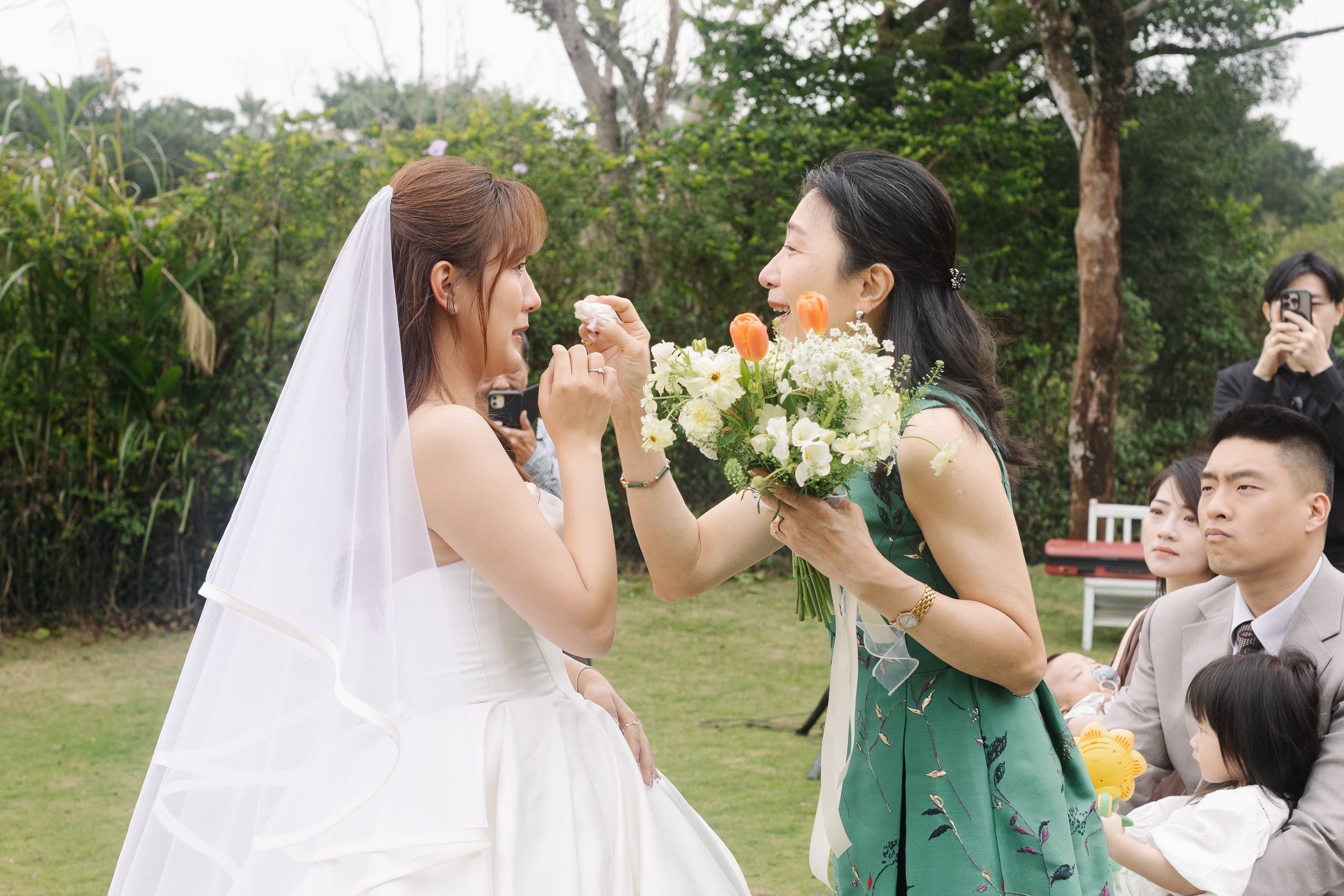 A bride and a woman exchanging a flower during a wedding ceremony outdoors, with guests watching and taking photos in the background.