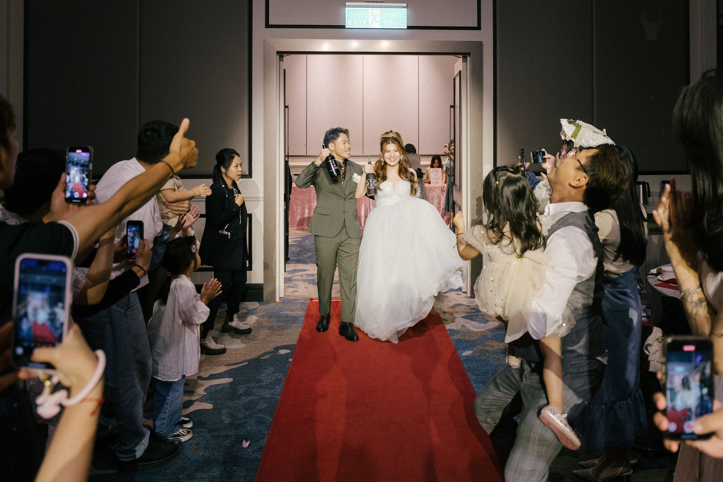 A bride and groom walking down a red carpet surrounded by guests taking photos at a wedding reception.