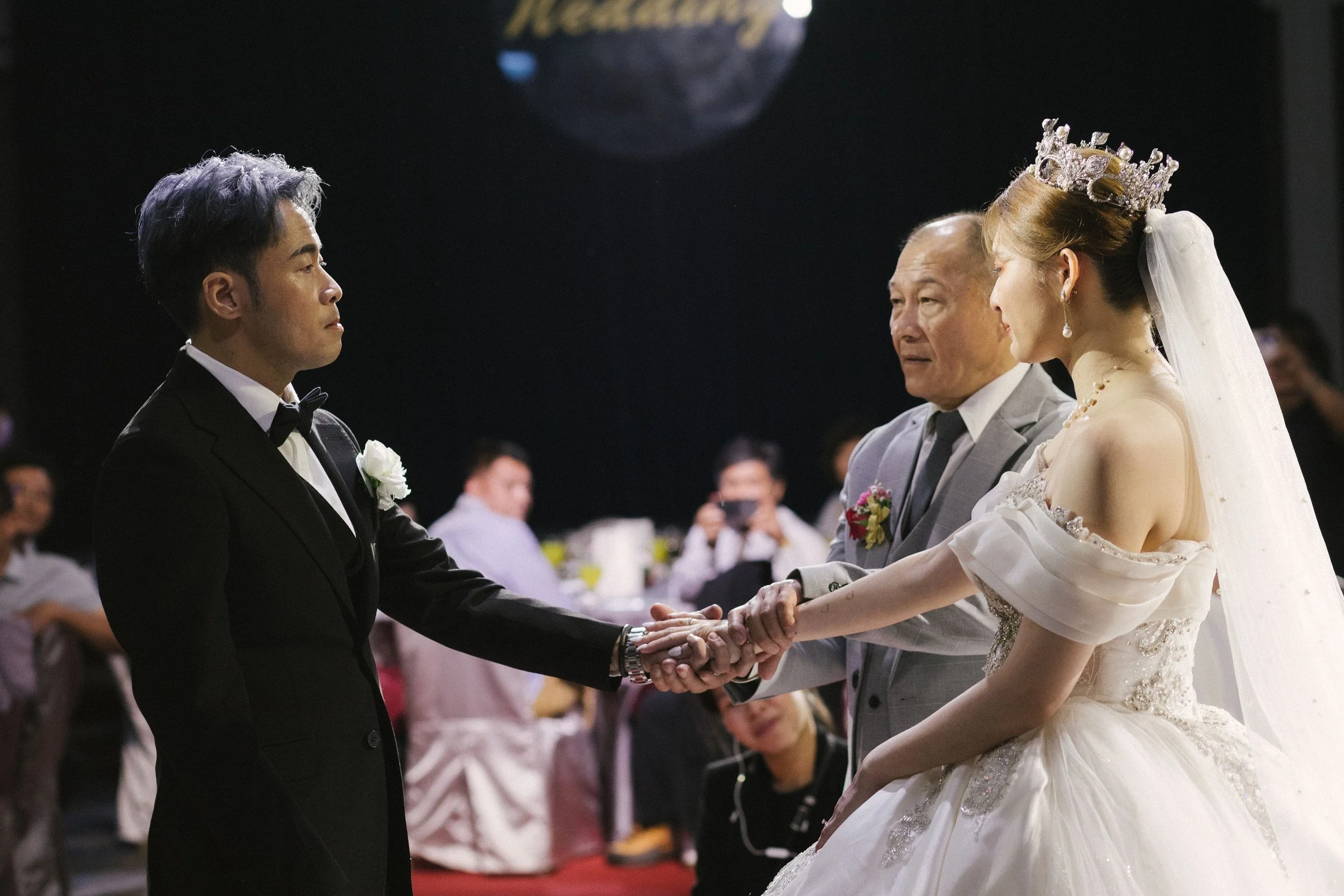 A wedding ceremony where a groom in a black tuxedo and a bride in a white wedding dress with a crown and veil hold hands with an older man in a gray suit. The groom looks serious, and the bride smiles softly. They are participating in a formal event 