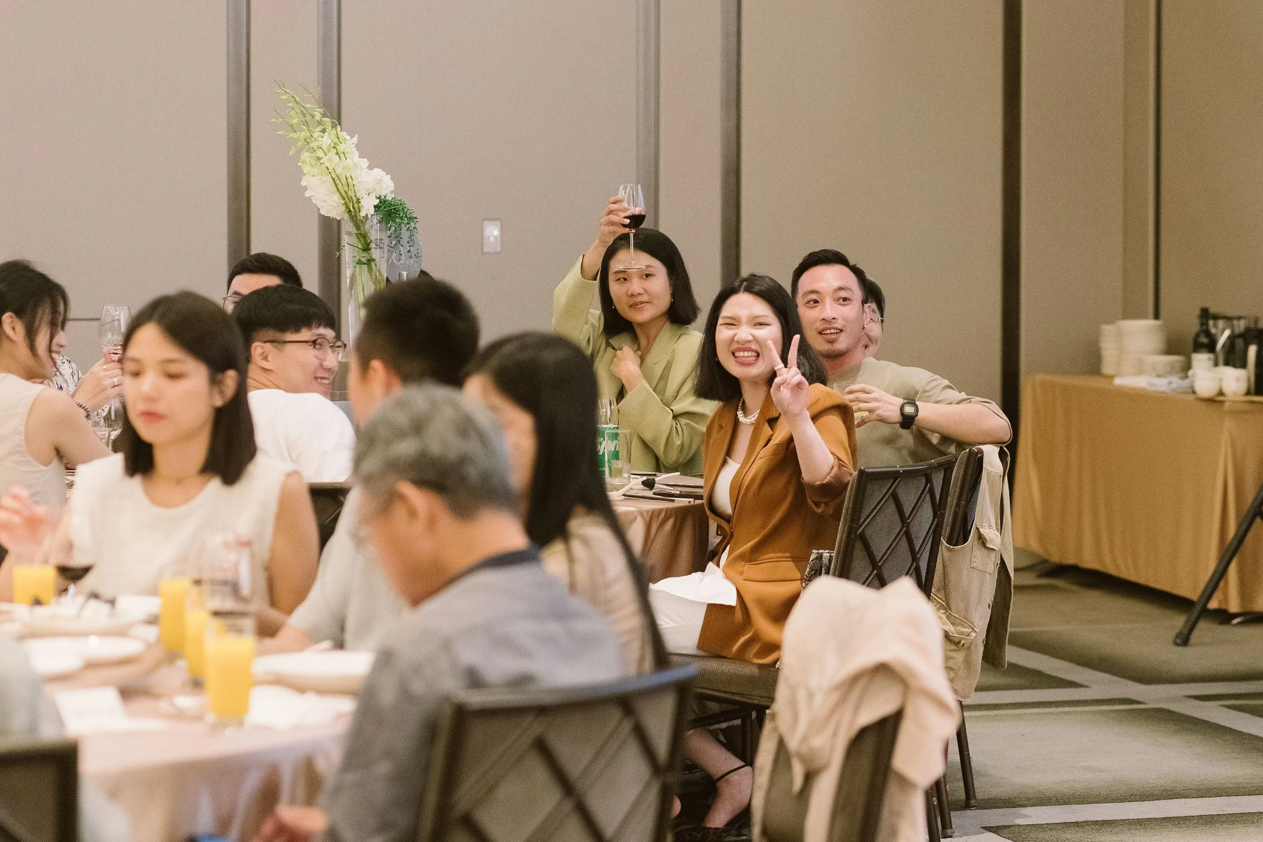 A group of people at a formal event seated around tables with food and drinks, with one woman holding up a glass of red wine and making a confused expression, and another woman making a peace sign and smiling.
