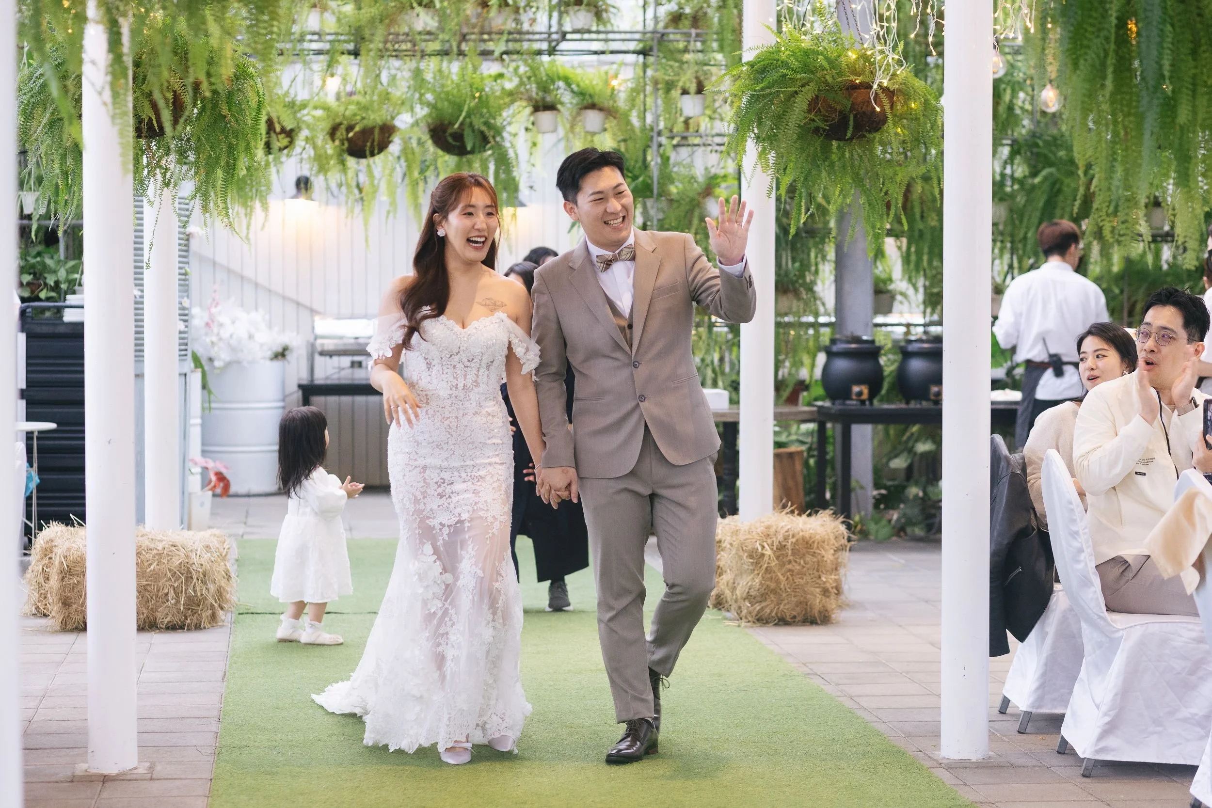 A newlywed couple walking hand in hand during their wedding reception, surrounded by green plants and hanging ferns in a decorated venue.