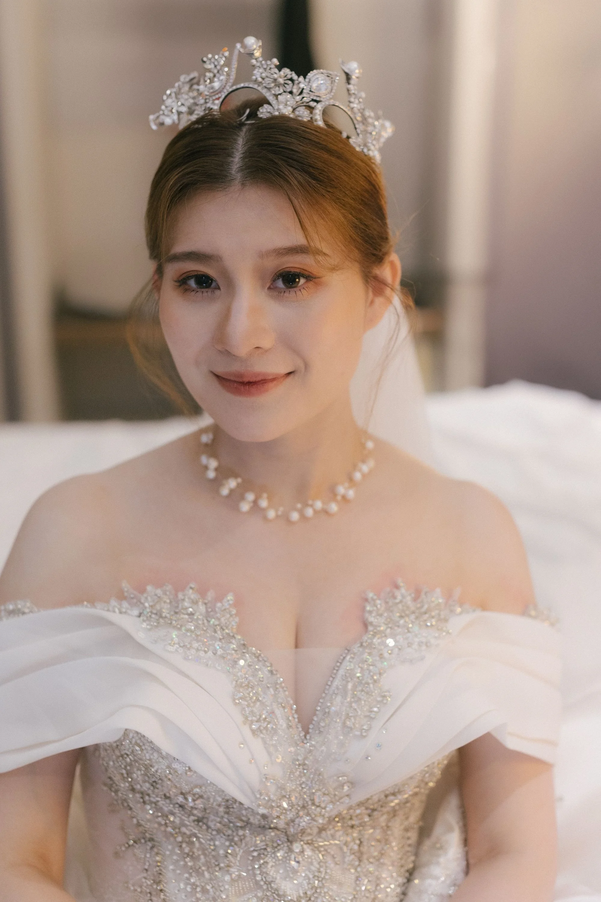 A bride wearing a sparkling white wedding gown with intricate beading and embroidery, and a pearl necklace, smiling softly.