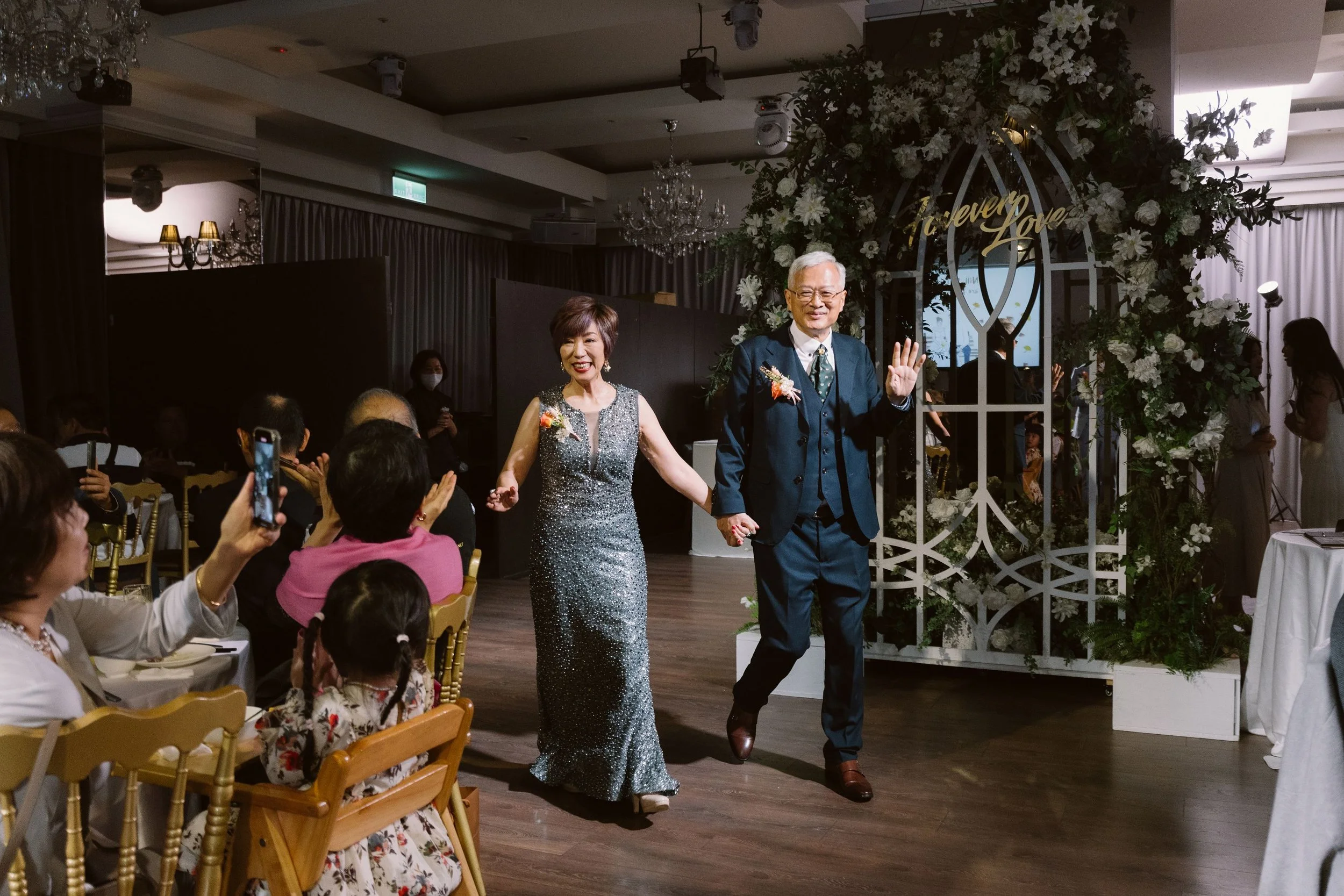 An elderly couple holding hands and smiling as they walk into a festive event with guests seated at tables and taking photos. The woman is wearing a silver, sparkly dress, and the man is in a dark suit with a boutonniere. There is floral decor and a 