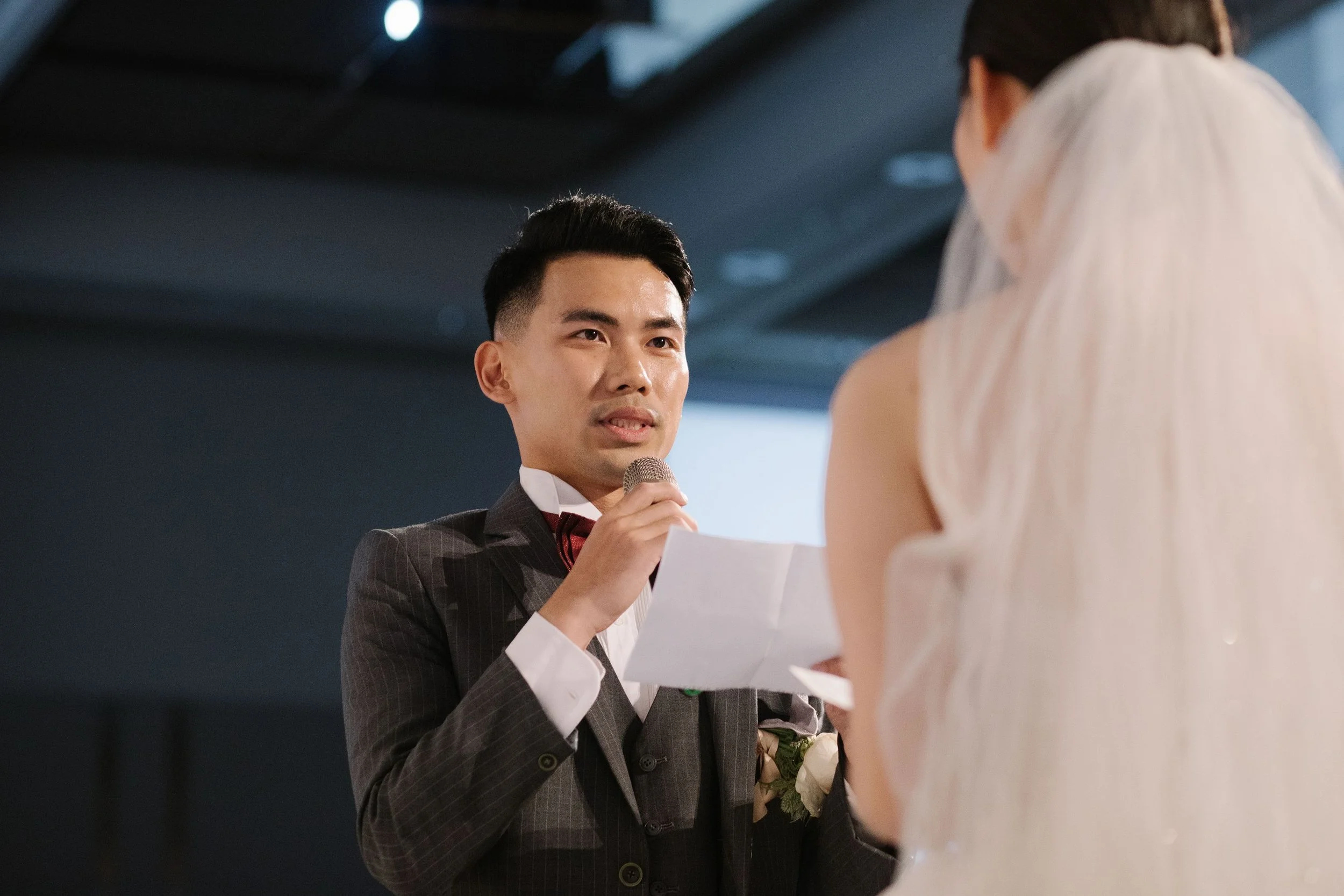 A man in a dark striped suit and burgundy bow tie with a white shirt is holding a microphone and reading from a paper to a woman in a light-colored dress, during a formal event or ceremony.