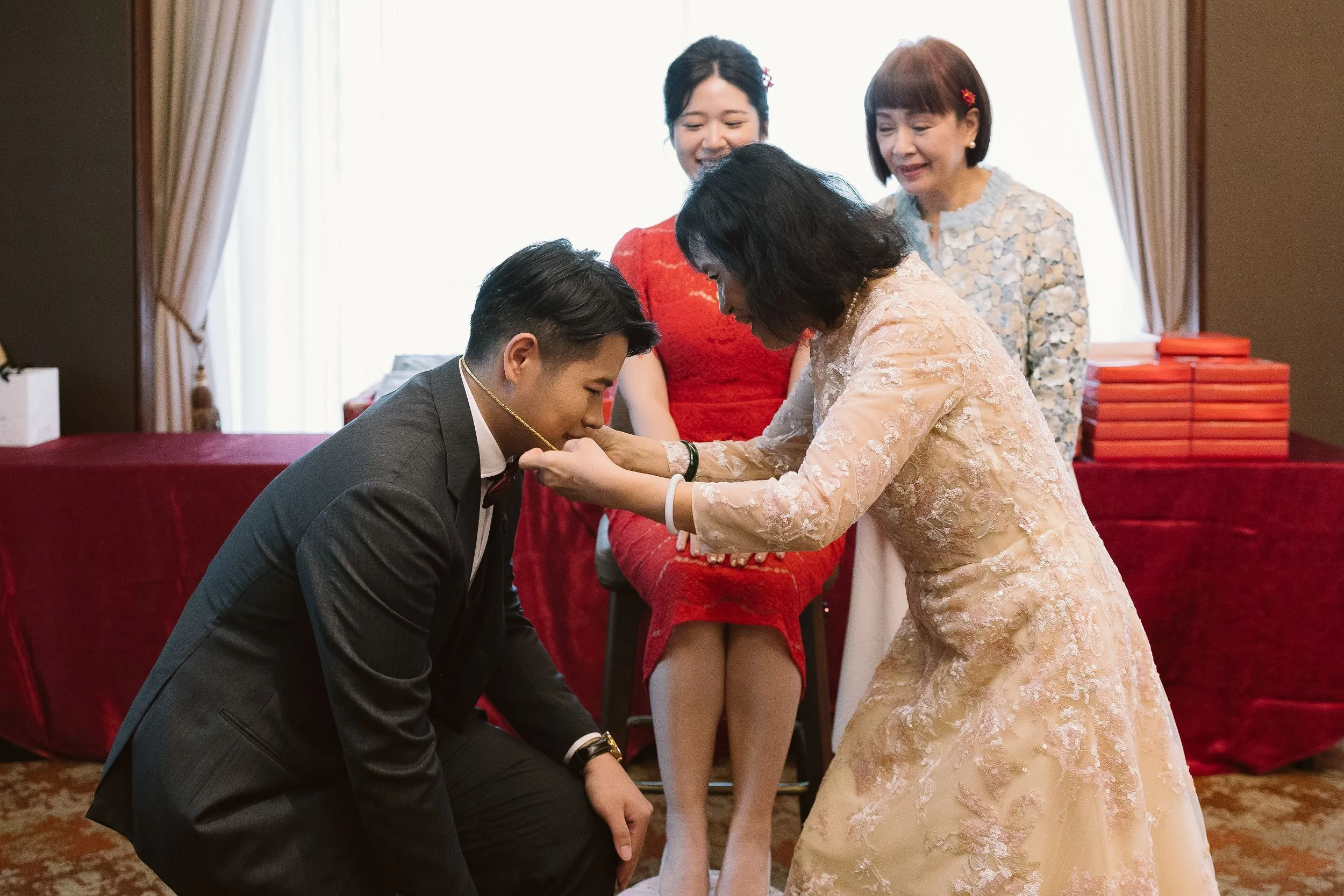 A woman in a peach lace dress bows her head as a man in a dark suit kisses her hand. Two women observe, one wearing a red dress and the other a patterned blouse, in a room with red drapes and a table with red wrapped gifts.