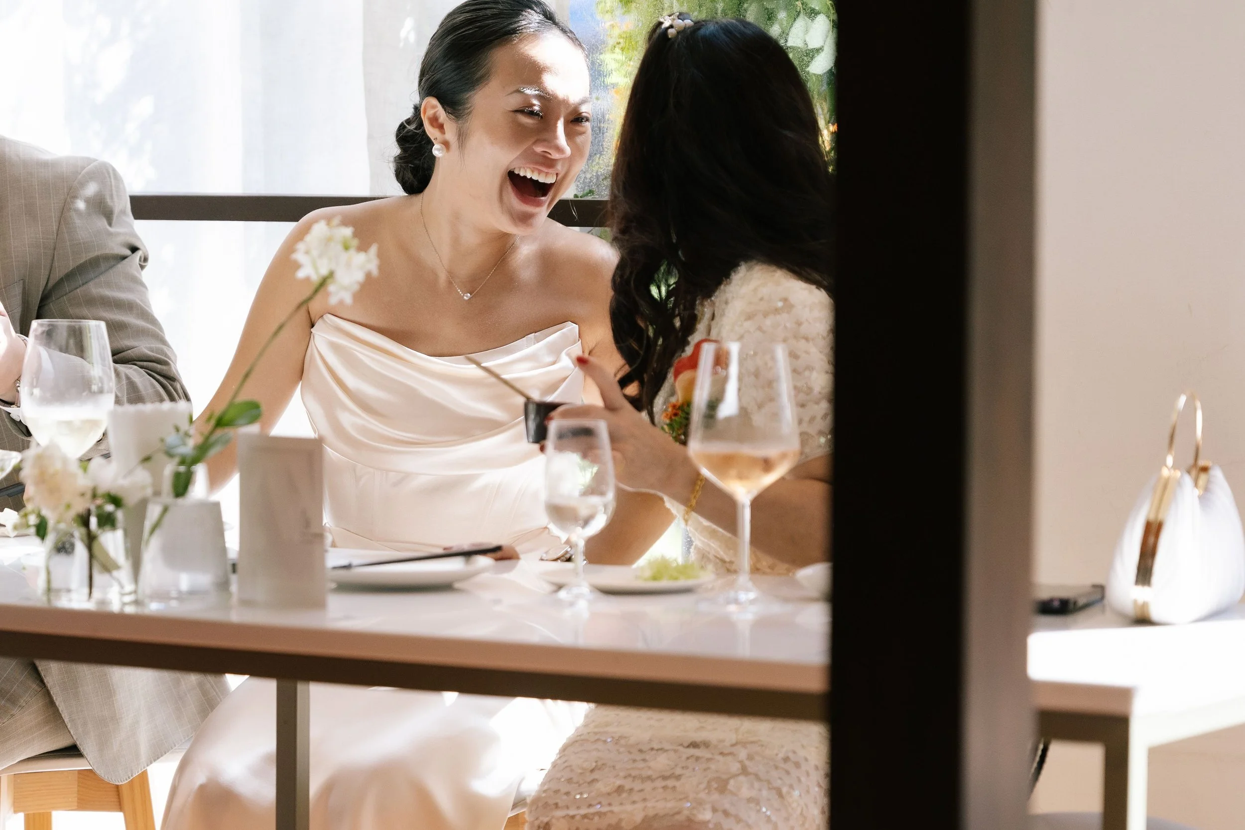 A woman in a white dress smiling and talking with another woman who has long dark hair, sitting at a table with glasses of wine and flowers in a bright room.
