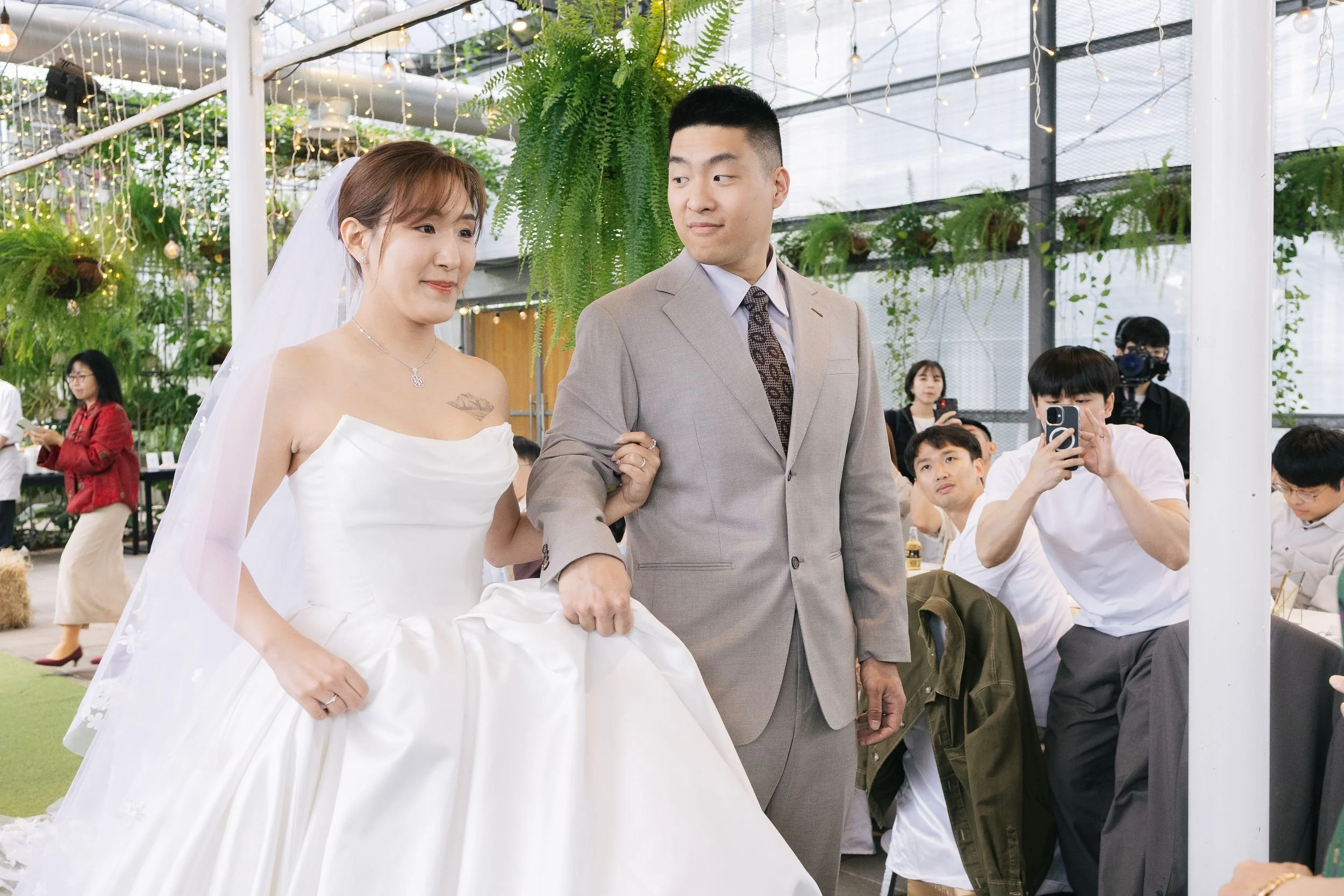 A bride in a white wedding gown with a veil and a necklace holding a man’s arm in a suit, at what appears to be a wedding reception or celebration indoors with green plants, hanging lights, and guests seated around.