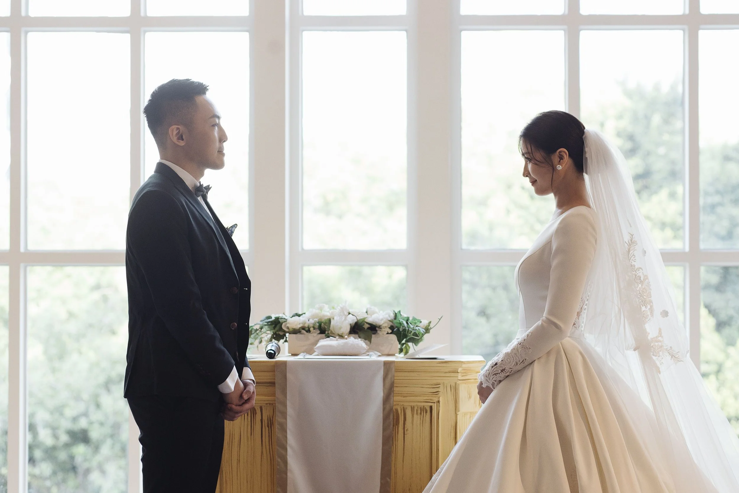 A bride and groom standing face to face during a wedding ceremony indoors with large windows in the background.