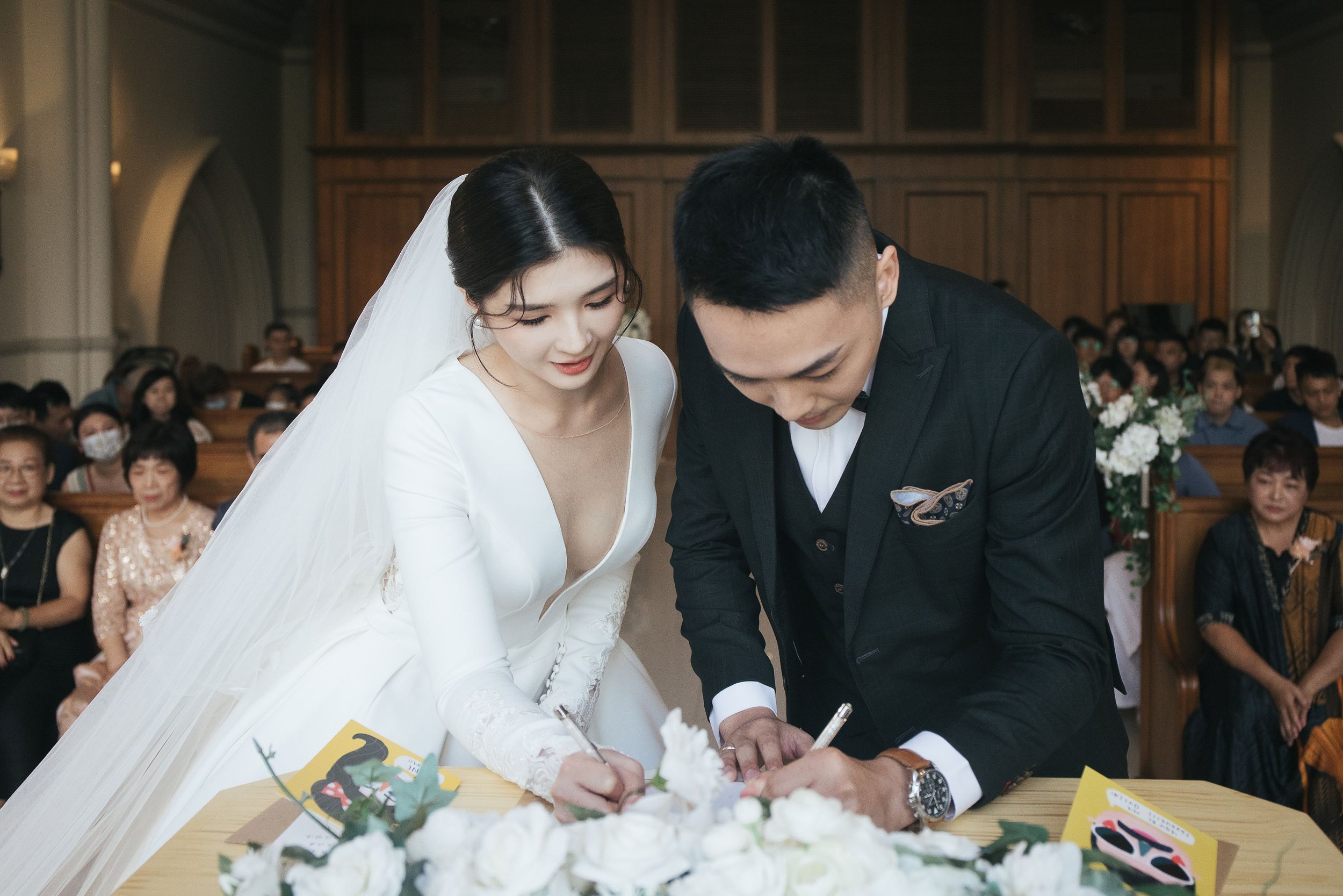 A bride and groom signing documents during their wedding ceremony in a church filled with seated guests.