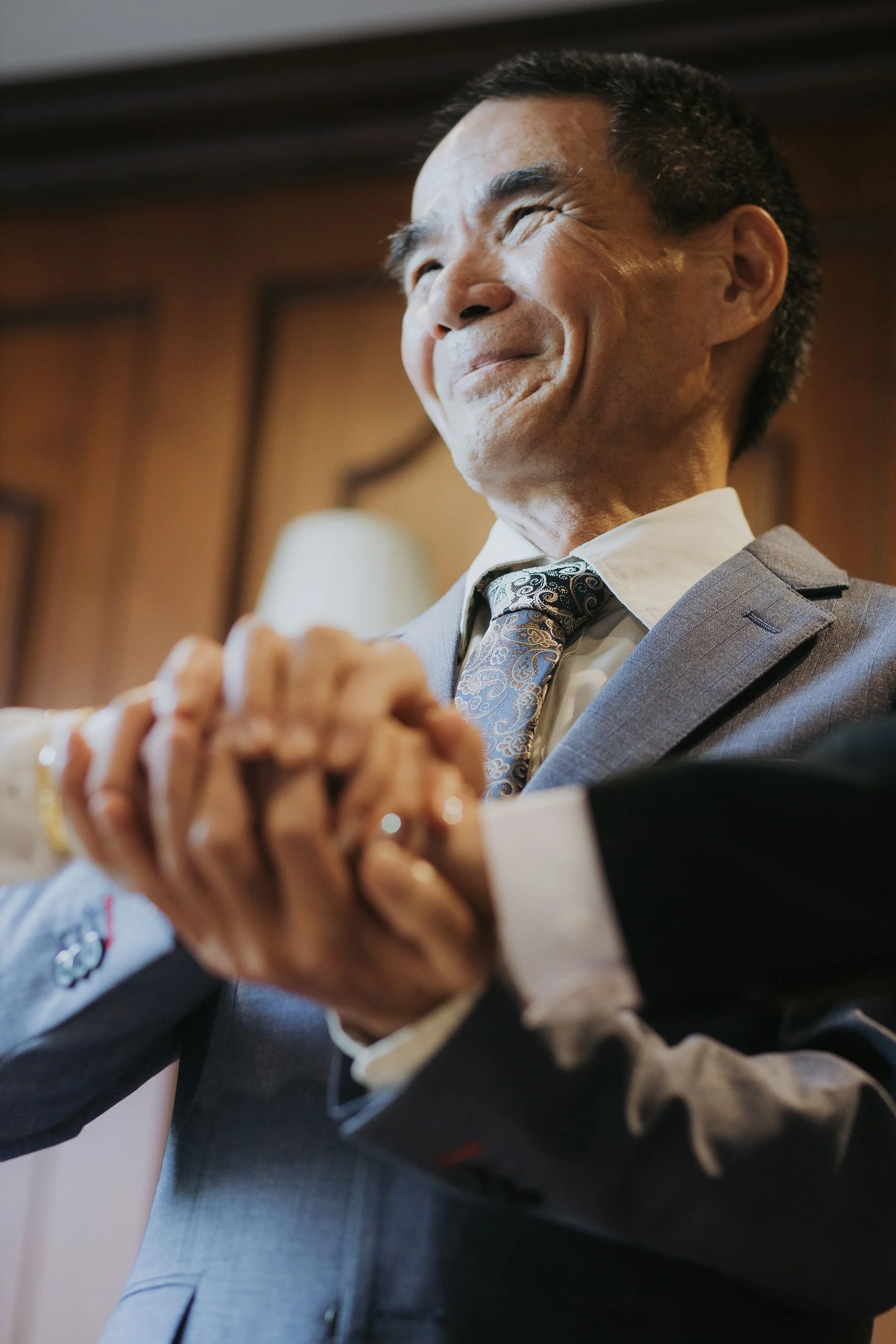 Close-up of an older man with a broad smile, holding hands with someone in front of him. He is wearing a gray suit, patterned tie, and a white dress shirt, with a warm indoor background.