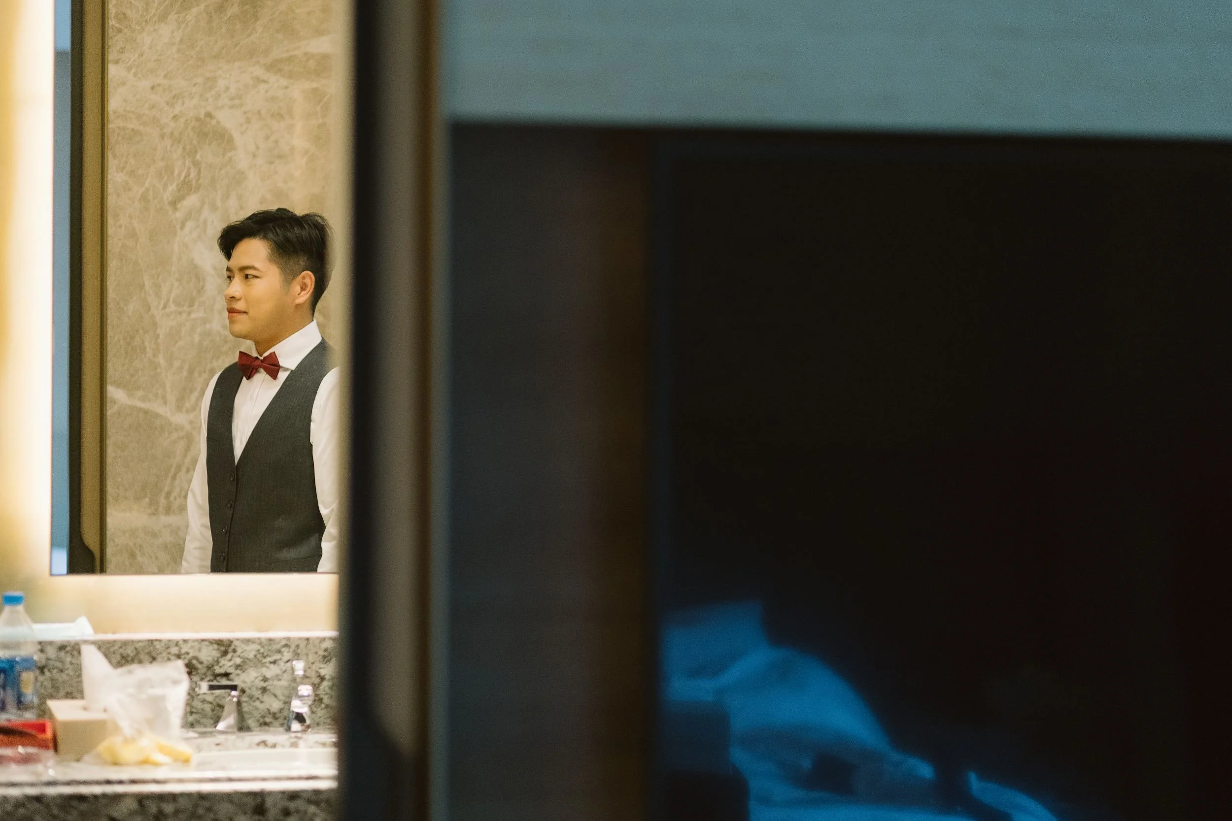 A man with dark hair dressed in a white shirt, black vest, and red bow tie seen through a mirror in a bathroom, with various toiletries on the counter.