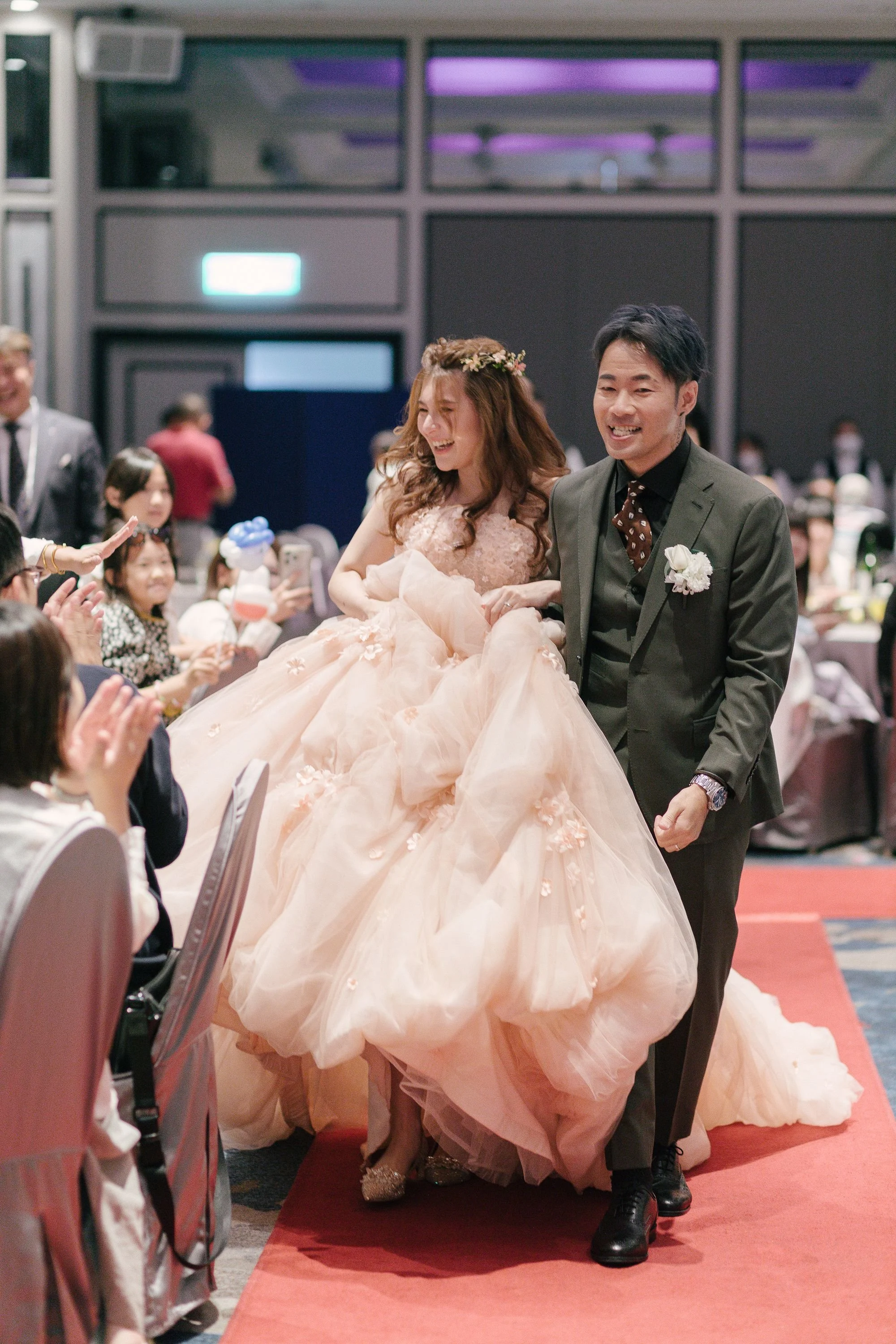 A bride in a peach-colored wedding gown with floral details, smiling and being led down a red carpet by a groom in a dark suit with a white flower boutonniere, at a wedding reception with seated guests and photographers taking pictures.