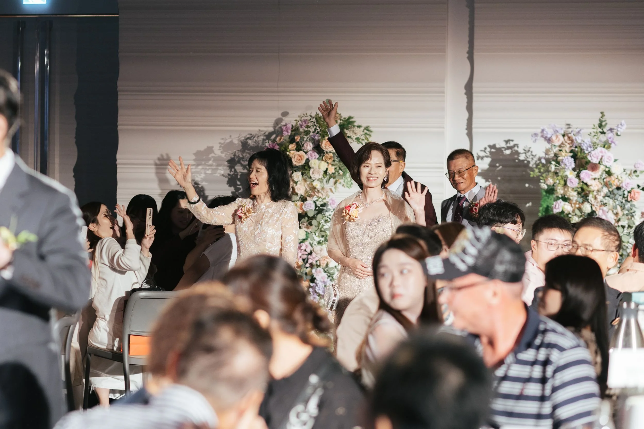 People at a wedding celebration, some smiling and waving, with floral decorations in the background.