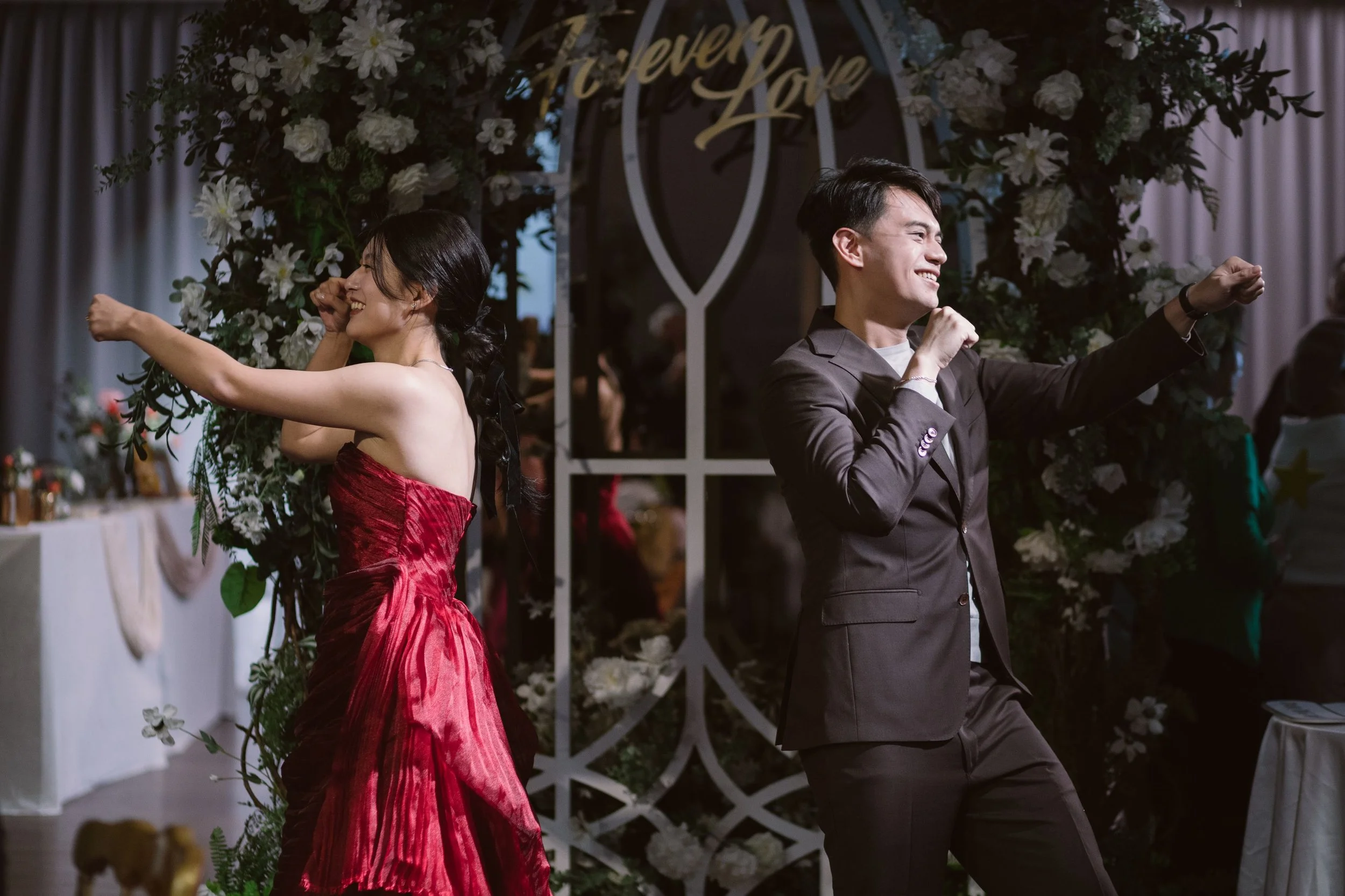 A woman in a red dress and a man in a suit are dancing back to back, smiling, at a wedding reception decorated with flowers and a sign that reads 'Forever Love'.
