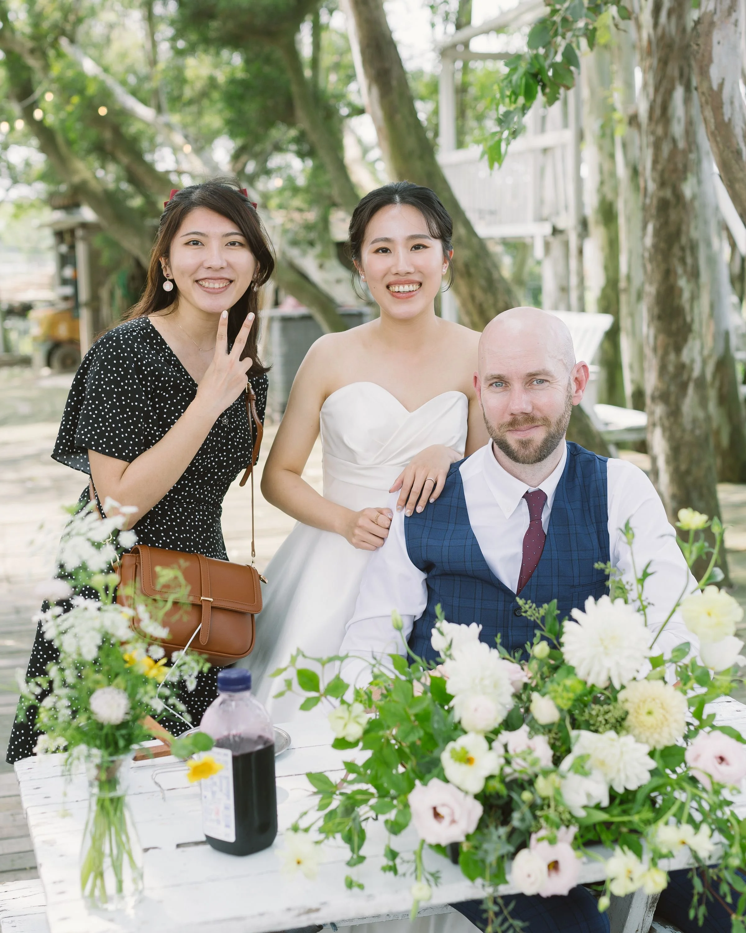Three smiling people, two women and one man, pose together outdoors at a garden or park during daytime. The woman on the left has long dark hair, wears a black polka dot dress, and makes a peace sign with her fingers. The woman in the middle is weari