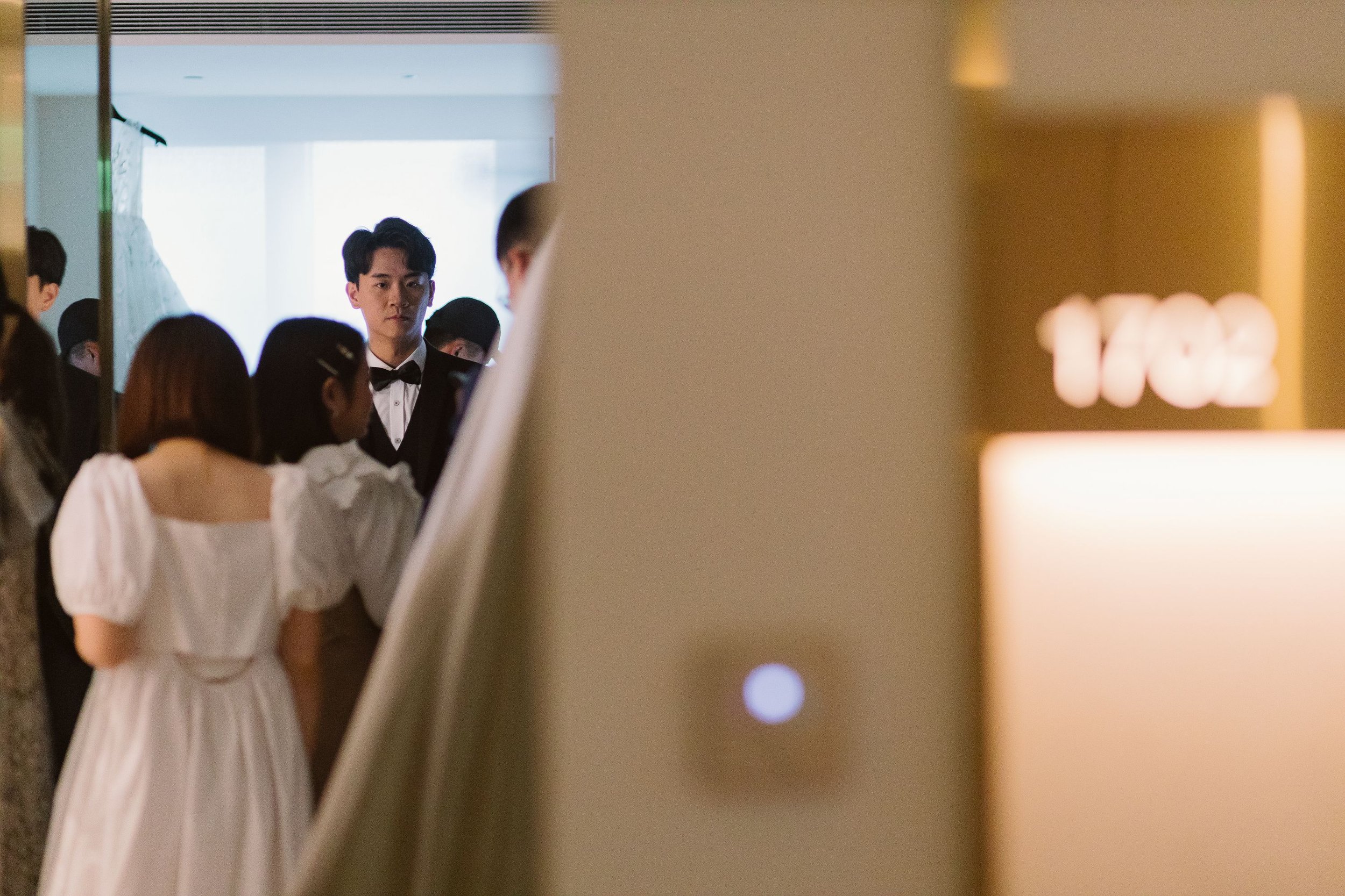 A group of people, including a young man in a tuxedo, gathered in a room with soft lighting, reflected in a mirror.