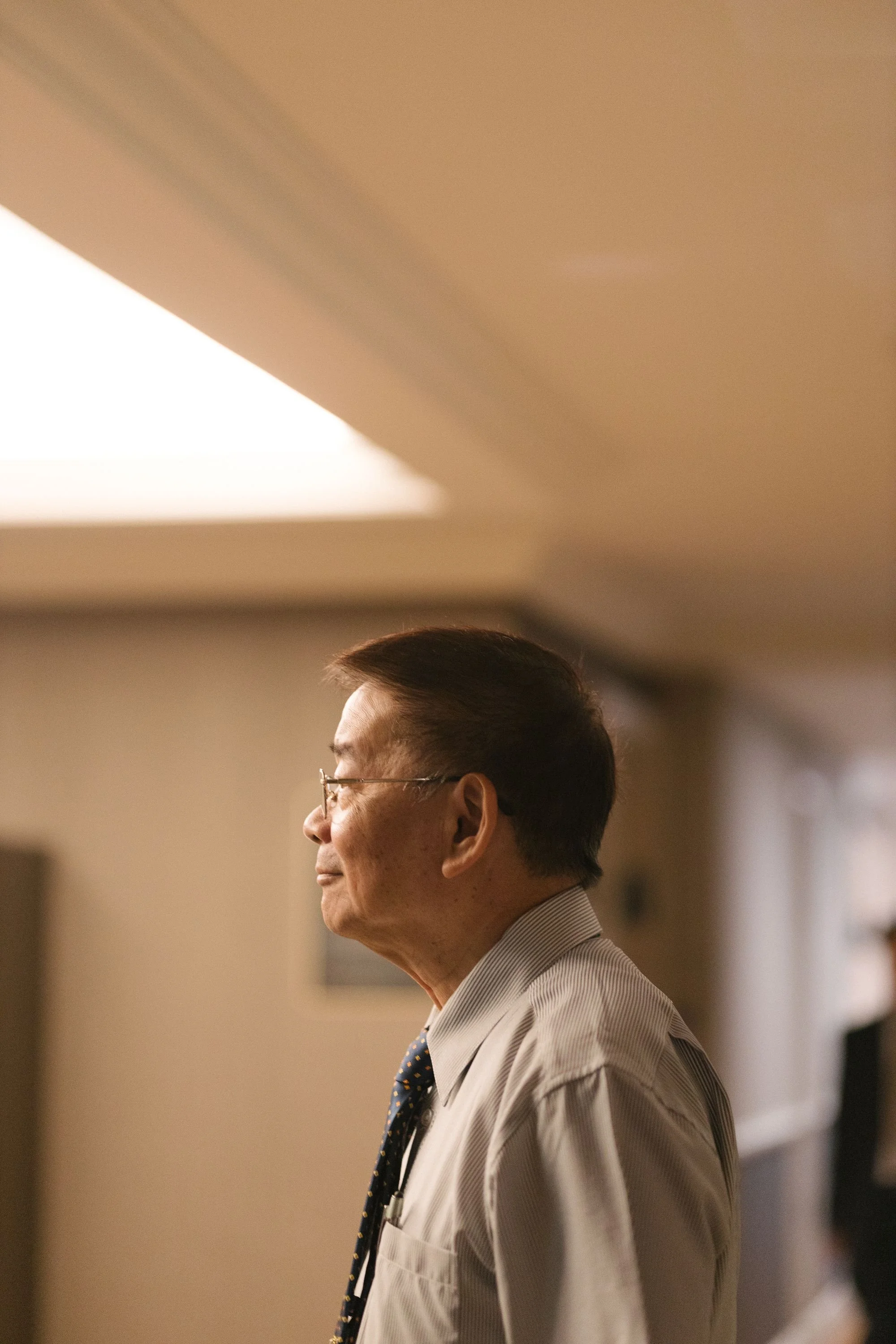 Side profile of an elderly man wearing glasses, a white shirt, and a patterned tie, standing indoors.
