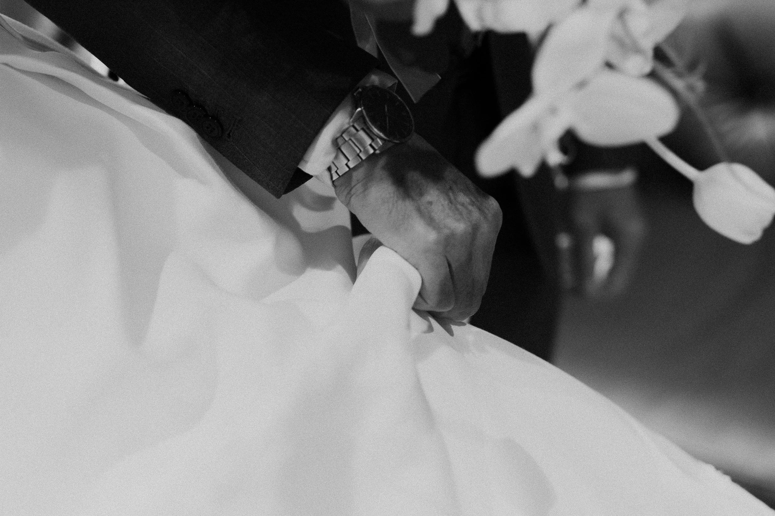 Close-up of a person wearing a suit and a wristwatch, with their hand resting on a white surface and a floral arrangement nearby, in black and white.