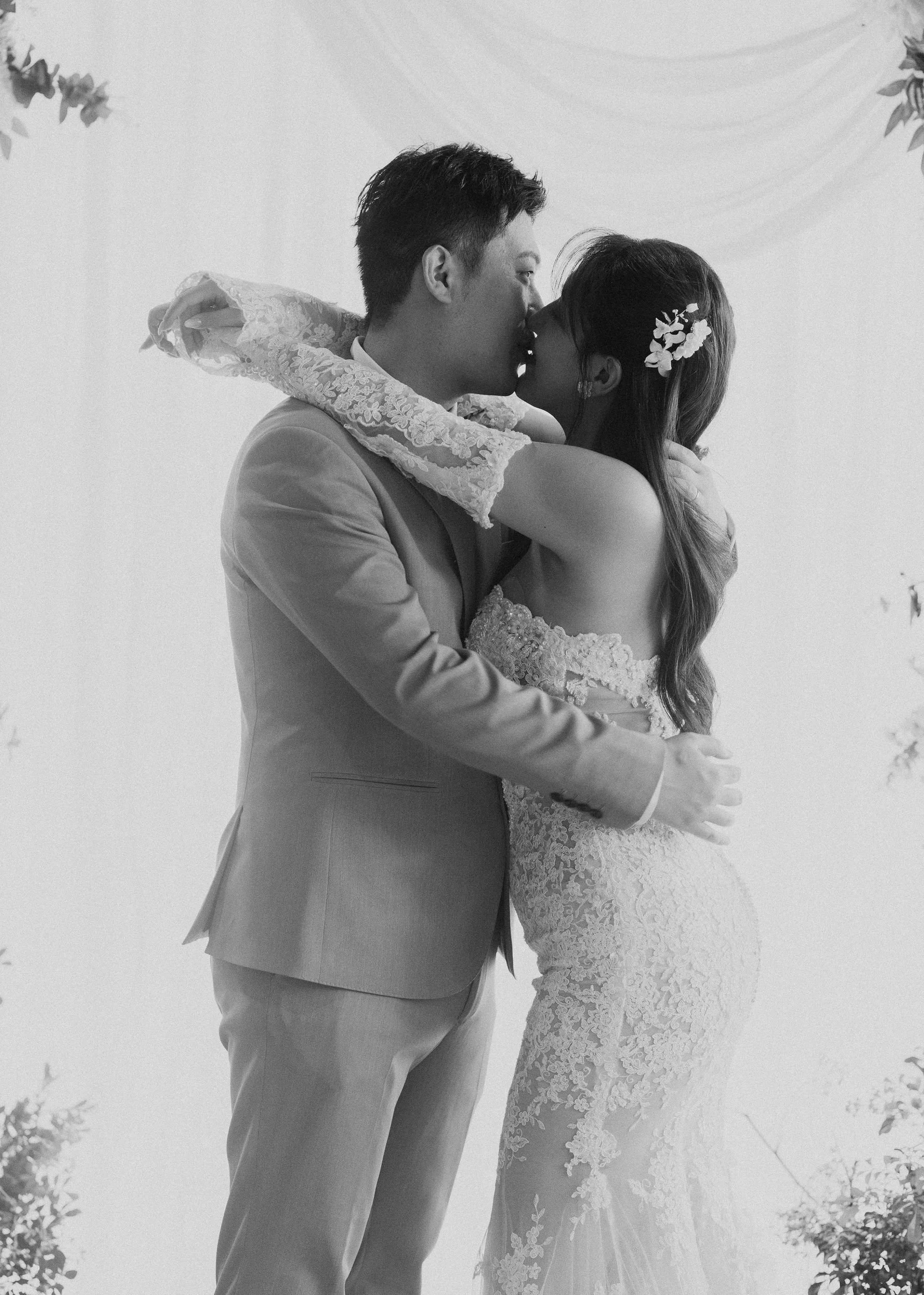 Black and white photo of a bride and groom kissing, with the bride's arms around the groom's neck, outdoors surrounded by trees.