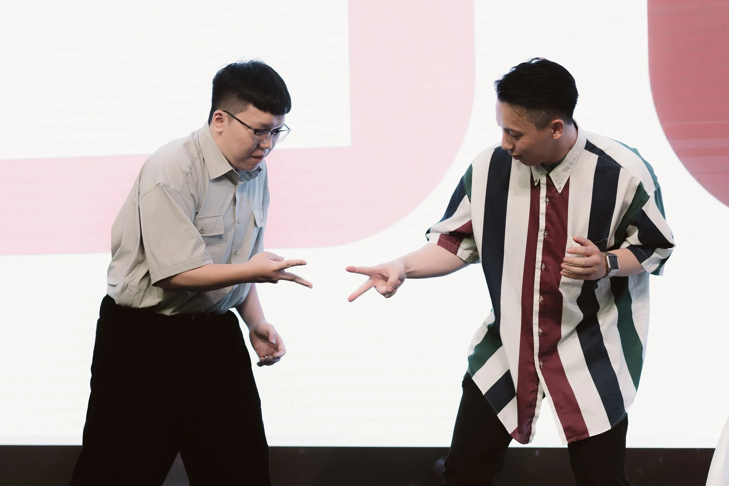 Two men with dark hair are standing facing each other, exchanging a small object between their hands. The man on the left is wearing glasses and a light-colored button-up shirt, while the man on the right is wearing a multicolored, striped button-up 