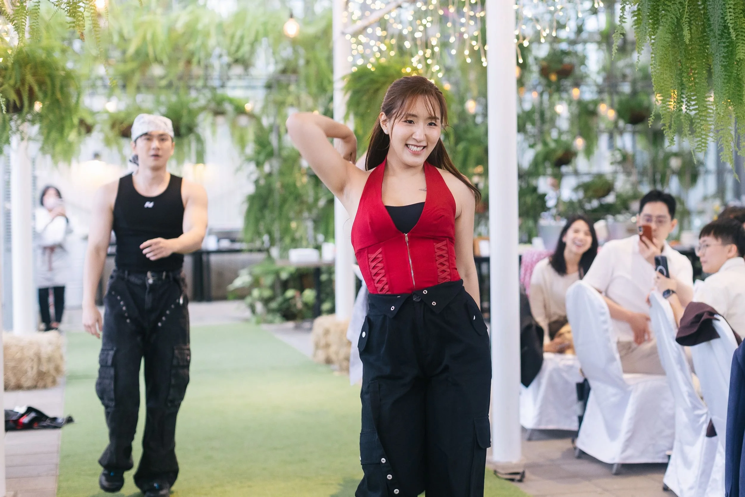 A young woman in a red top and black pants is smiling and posing with her hand behind her head. She is in an indoor setting with greenery and hanging lights. Several other people are in the background, including a man in a black tank top and head sca