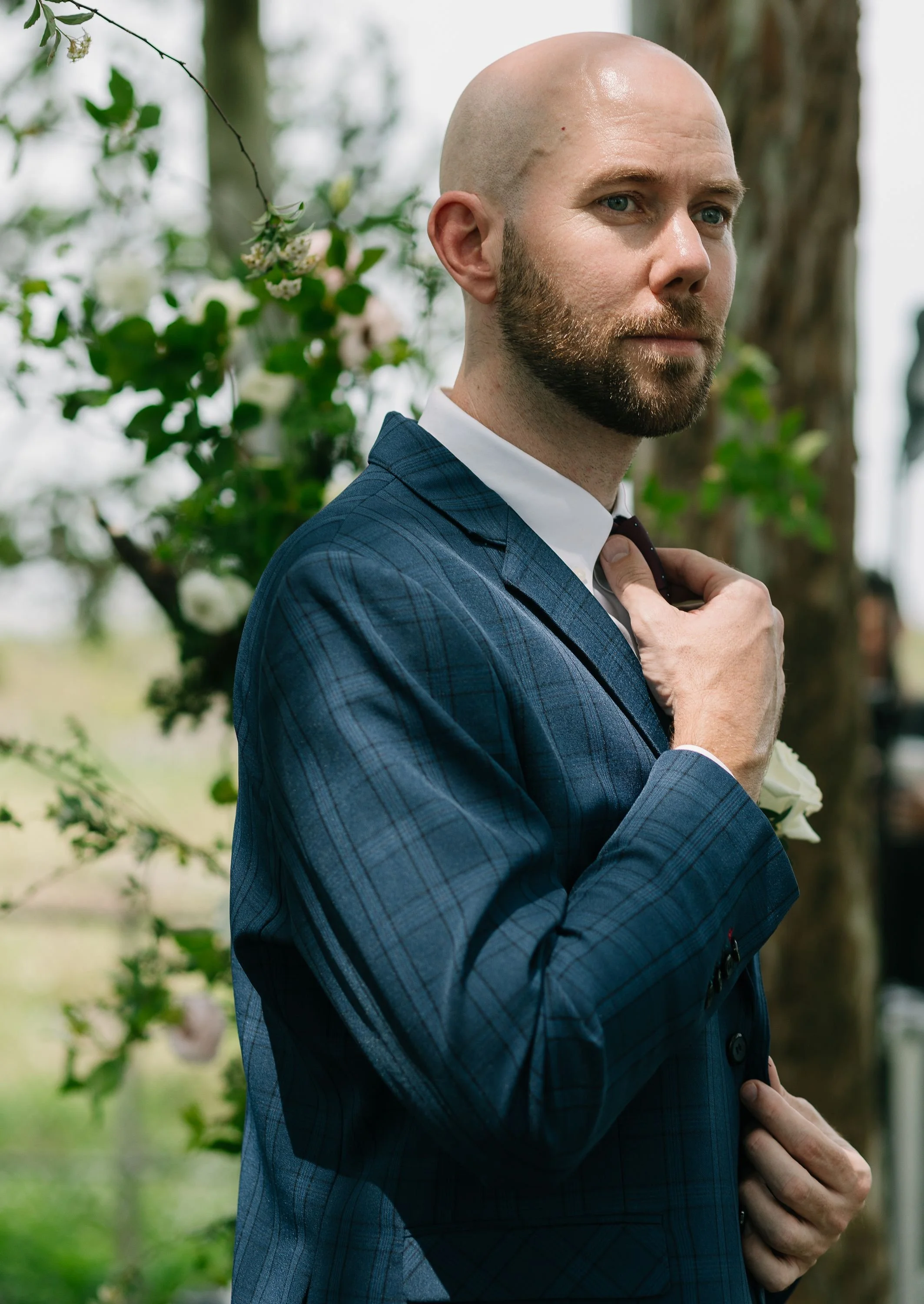 A man with a bald head and beard adjusting his tie while standing outdoors with greenery and flowers in the background.
