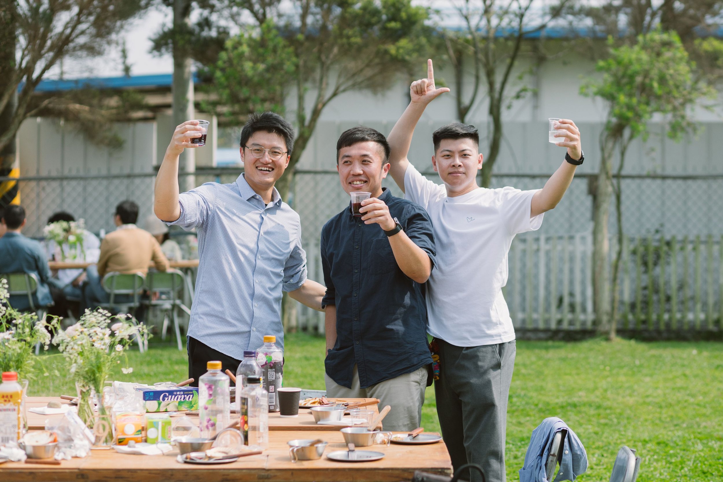 Three young men standing outdoors, smiling and holding drinks, with a table of food and drinks in front of them and other people sitting at tables in the background.