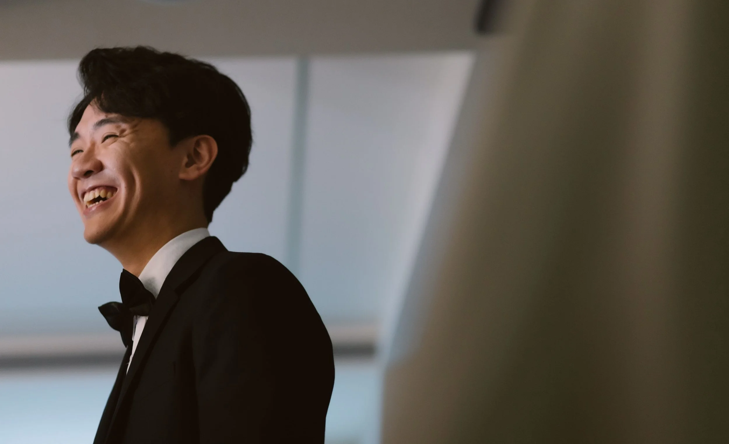A man in a tuxedo with a bowtie, smiling and showing teeth, standing in an indoor setting.