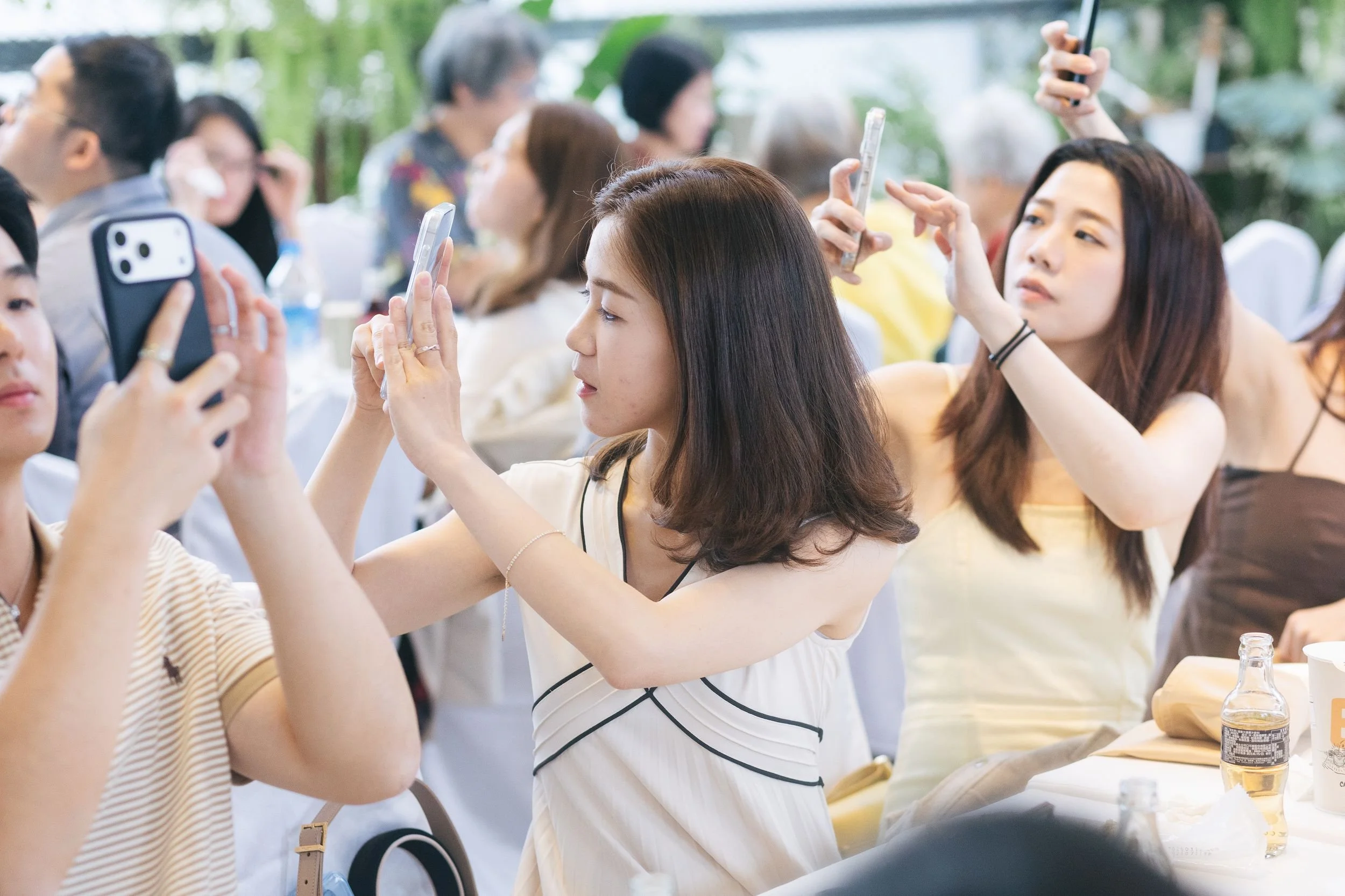 A group of women sitting at a table, taking photos with their smartphones, at an outdoor event surrounded by greenery.